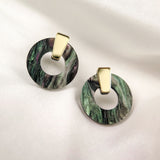 A pair of round, marbled green and black acrylic glass earrings with gold triangular studs, displayed on a soft white fabric background. These hypoallergenic Minimalist Circle Stud Earrings combine style and comfort beautifully.