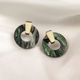 Minimalist Circle Stud Earrings featuring gold geometric studs and circular, marbled green and black hoops crafted from acrylic glass, displayed on a white fabric background. Hypoallergenic for comfortable wear.