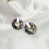 A pair of round, black Minimalist Circle Stud Earrings with colorful iridescent flecks and gold rectangular studs, crafted from hypoallergenic acrylic glass, displayed on white satin fabric.