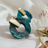 Two minimalist circle stud earrings with a marbled teal design and small gold accent, crafted from hypoallergenic acrylic glass, are displayed on a white cloth next to a white flower with pink details.