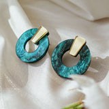 A pair of minimalist circle stud earrings crafted from teal marbled acrylic glass with gold geometric accents rest on shiny white fabric, with a blurred white flower in the foreground. Hypoallergenic for sensitive ears.