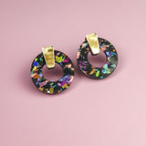 A pair of minimalist circle stud earrings with multicolored, iridescent patterns crafted from acrylic glass and gold rectangular clips, displayed on a pink background.