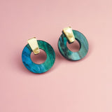 Minimalist Circle Stud Earrings crafted from blue-green marbled acrylic glass with gold triangular tops, displayed on a pink background. These hypoallergenic earrings offer both style and comfort.
