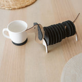 A wooden dachshund-shaped coaster holder with black round felt coasters on its body sits on a light wooden table next to a white mug, with woven placemats in the background.