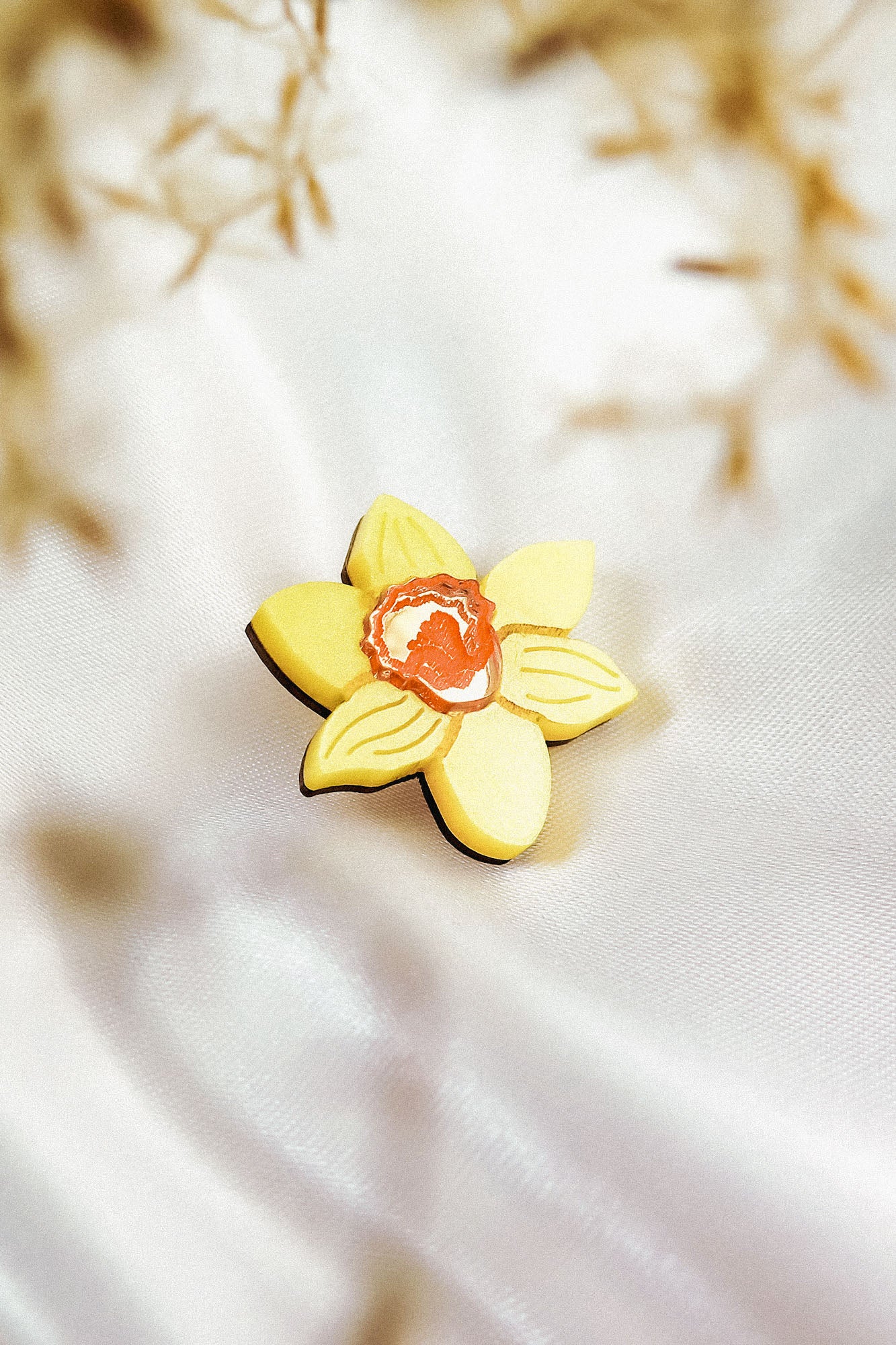 A yellow, handmade flower-shaped cookie with an orange center rests on white, silky fabric, resembling a Daffodil Flower Lapel Pin, with blurred brownish plants in the background.