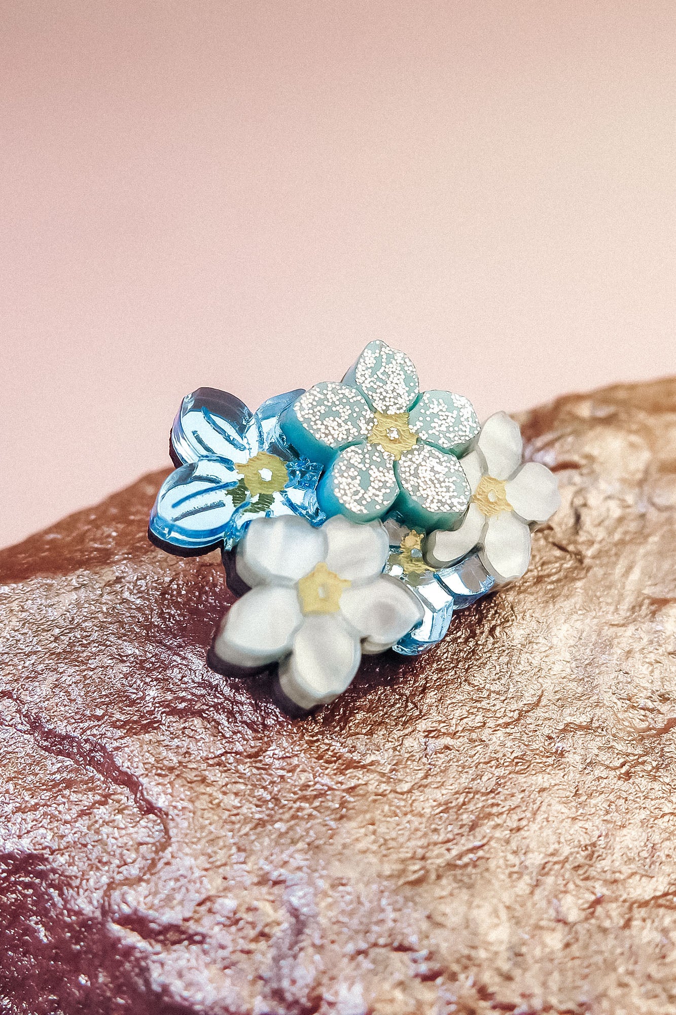 A cluster of delicate, handmade Forget Me Not Flower lapel pins with pastel blue and white petals and yellow centers rests on a textured rock against a soft pink background.