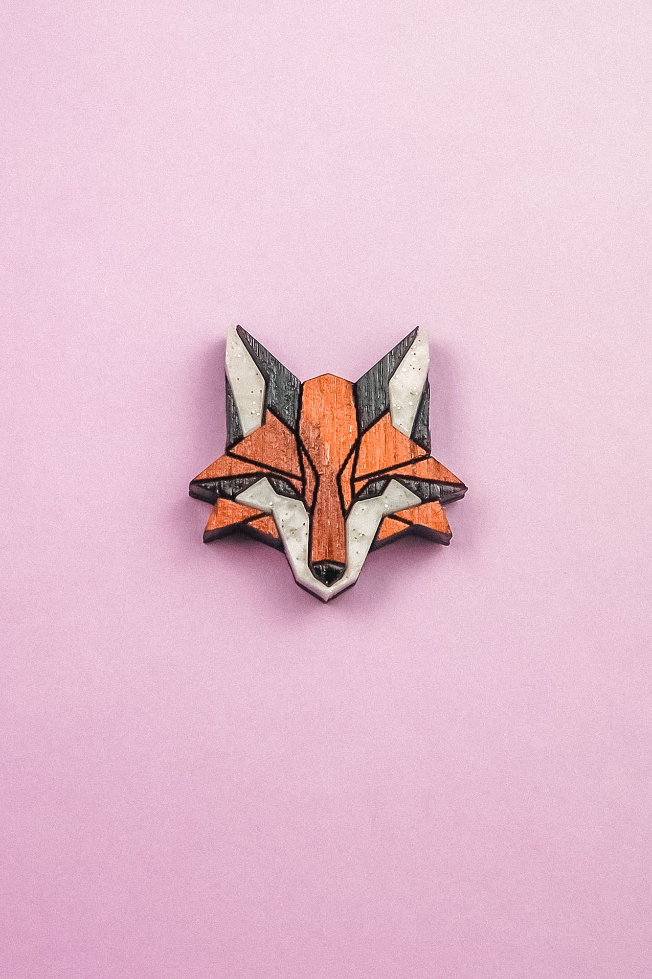 A geometric Fox Pin brooch, crafted from recycled wood in orange, black, and white hues, is displayed on a pink background—perfect for an eco-conscious style.