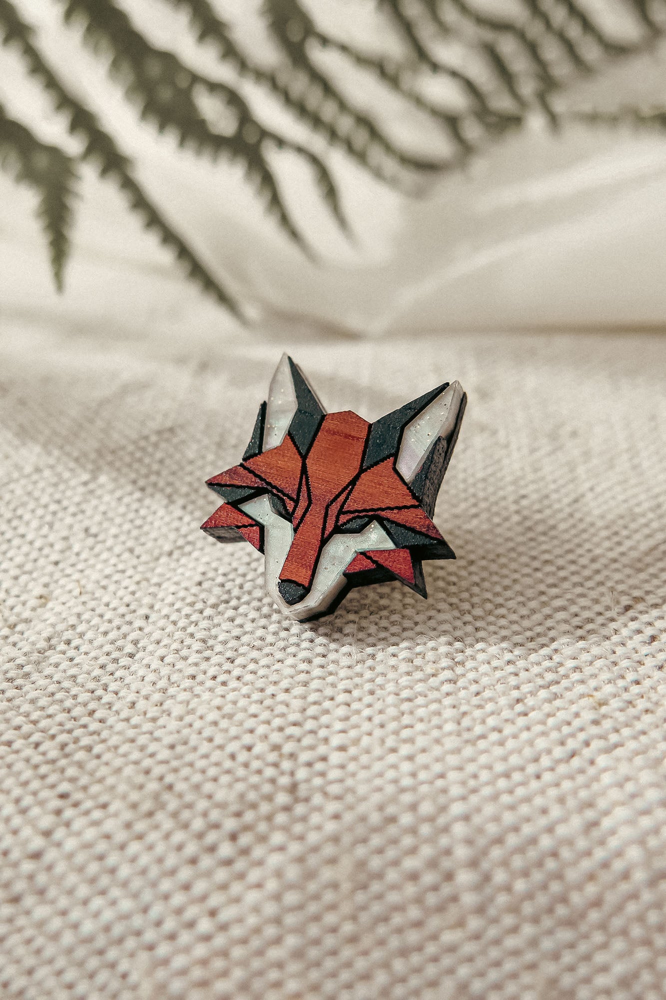 A geometric Fox Pin crafted from recycled wood in red, black, and white rests on textured cream fabric, with blurred green fern leaves adding an eco-conscious style to the scene.