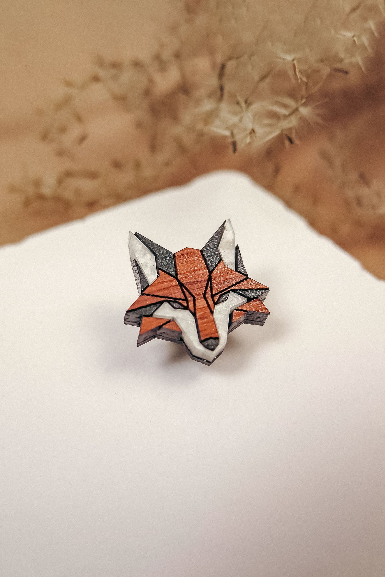 A geometric Fox Pin brooch, crafted from recycled wood in orange, black, and white tones, rests on a white surface with blurred dried plants in the background—a perfect touch for an eco-conscious style.