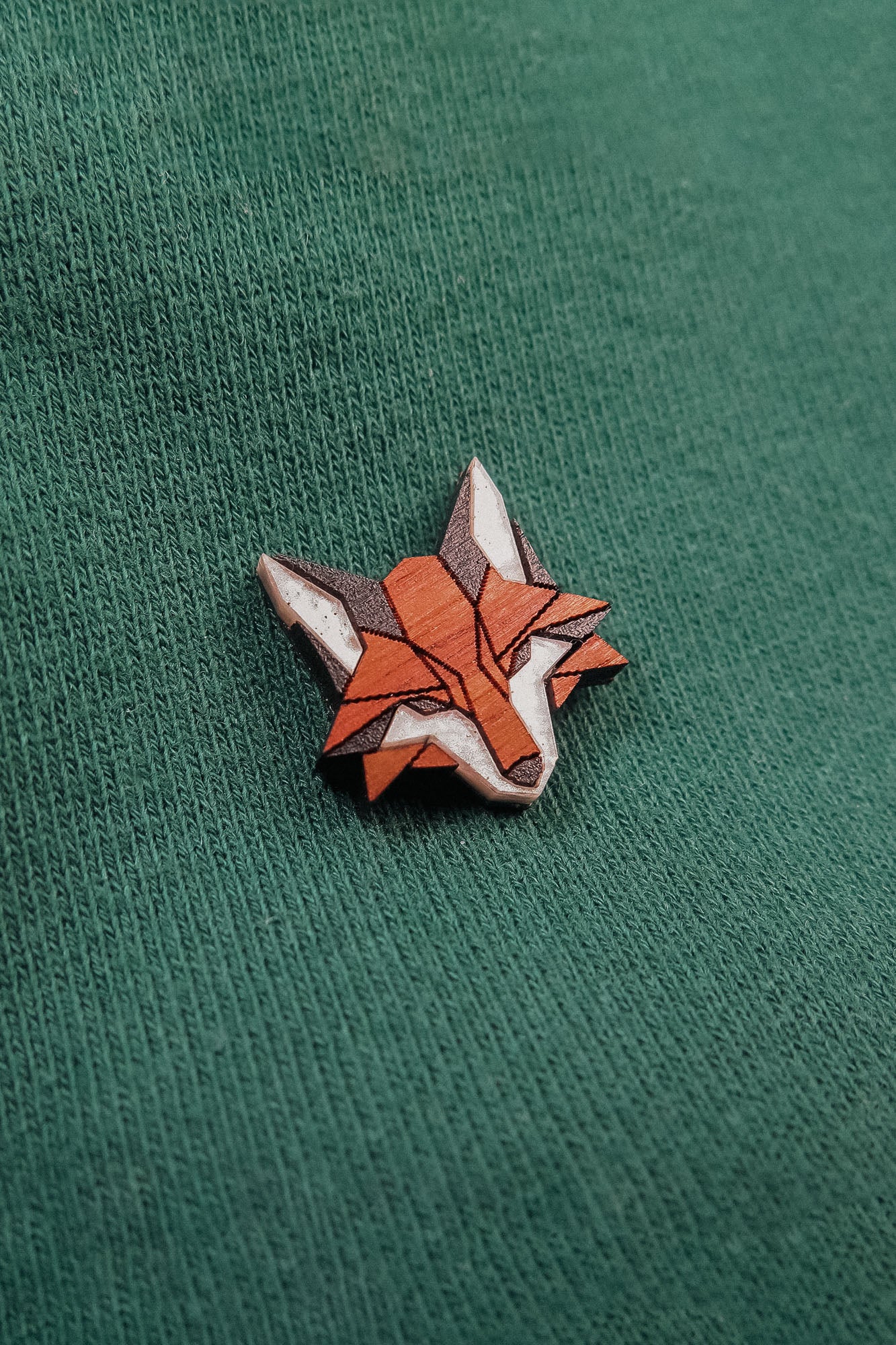 A geometric Fox Pin crafted from recycled wood, featuring orange, white, and brown details, is attached to a green fabric background—perfect for showcasing your eco-conscious style.