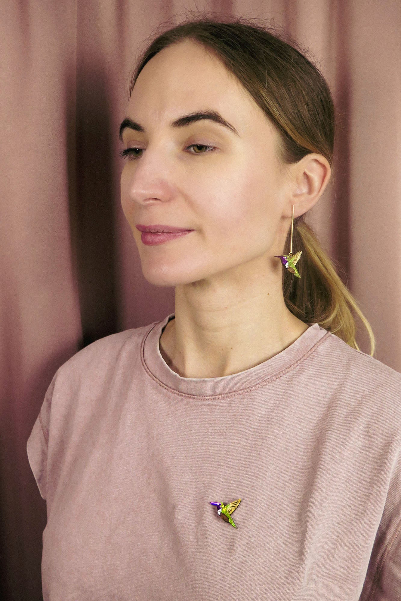A woman with light brown hair in a ponytail wears a light pink shirt, a colorful hummingbird earring, and a nature-inspired hummingbird pin while standing in front of a pinkish curtain.