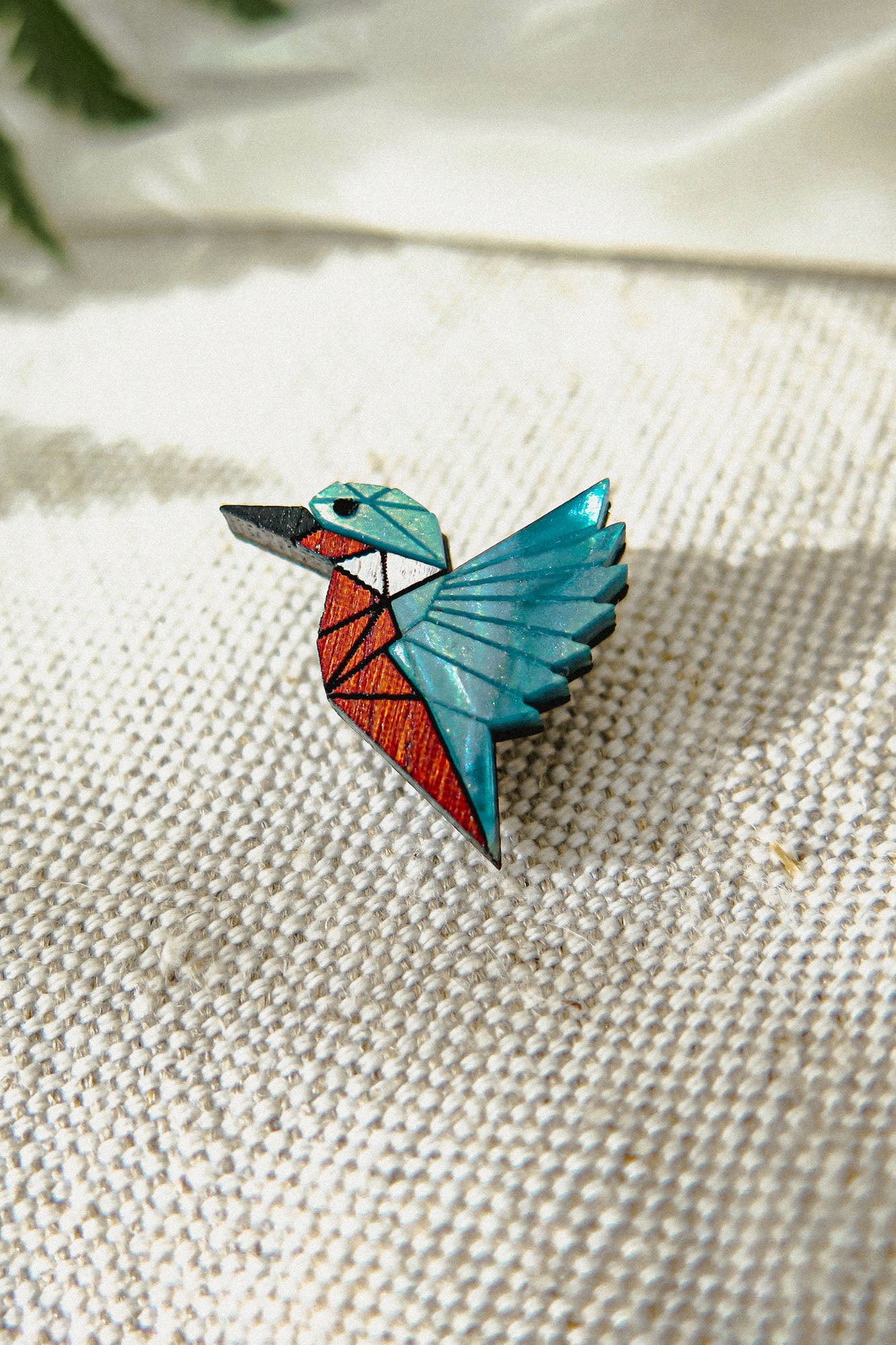 A kingfisher pin brooch shaped like a bird with geometric patterns in teal, red, black, and brown, crafted from recycled wood and placed on textured beige fabric with soft natural light.