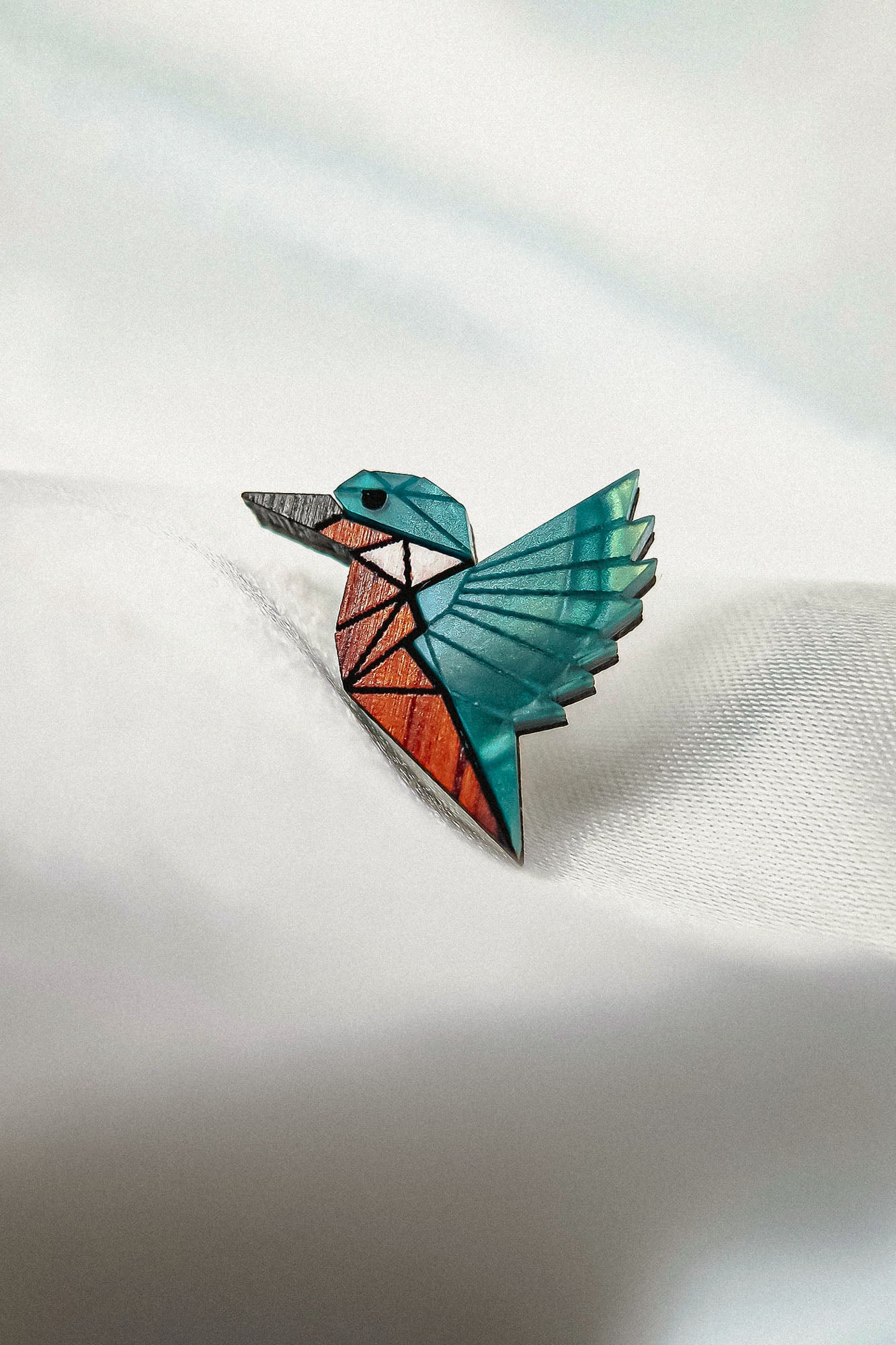 A colorful, geometric brooch shaped like a hummingbird rests on a white, softly textured surface. Crafted from eco-friendly materials, the brooch features teal, orange, and brown sections with black outlines in a stained-glass effect.