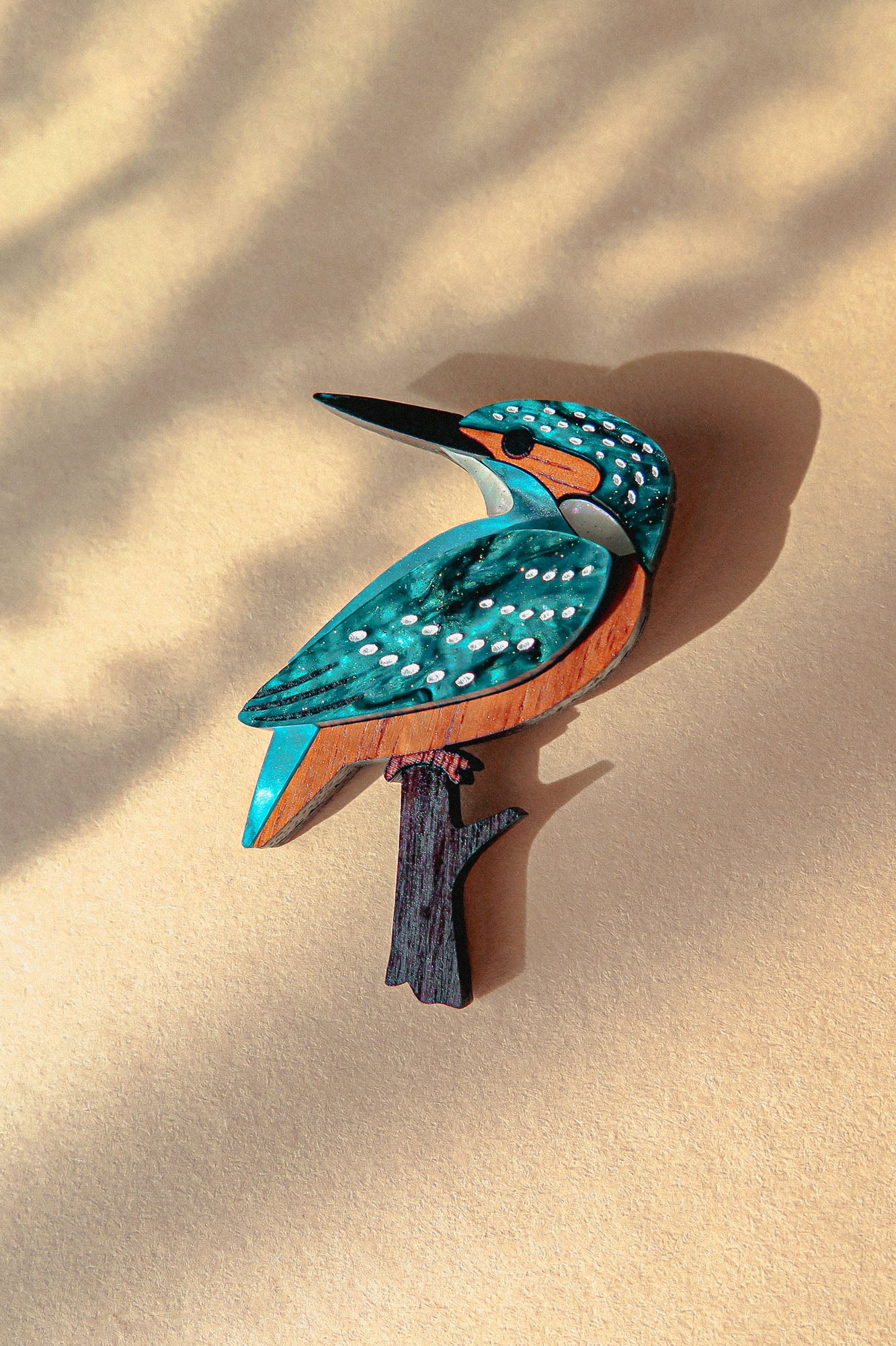 A colorful, handcrafted Kingfisher Brooch made from sustainable materials rests on a beige surface with soft shadows. The wooden brooch features striking blue, teal, white, and brown details.