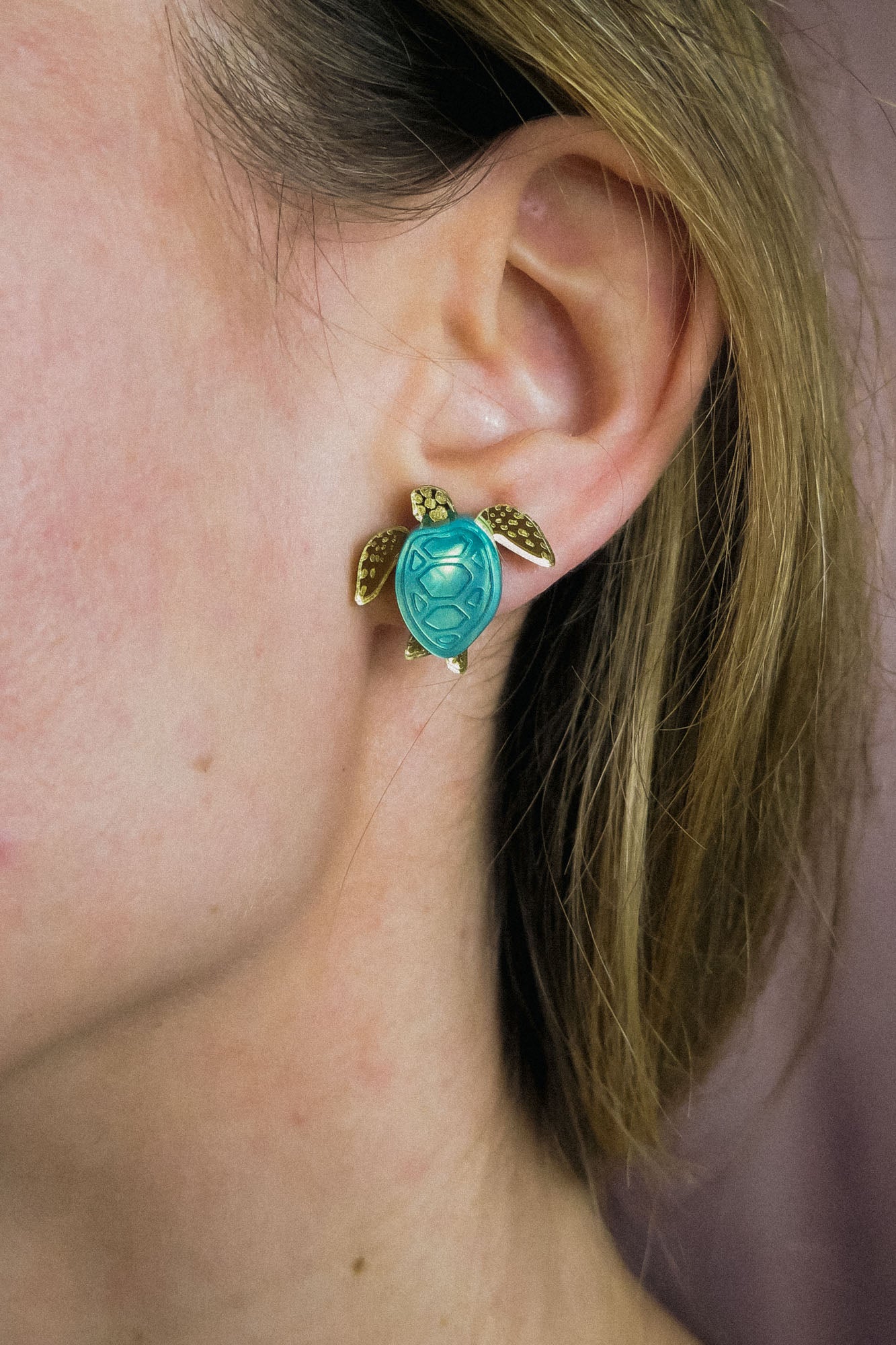 A close-up of a persons ear wearing gold and turquoise Sea Turtle Studs, with their light brown hair partially visible, captures an ocean-inspired, summery vibe.