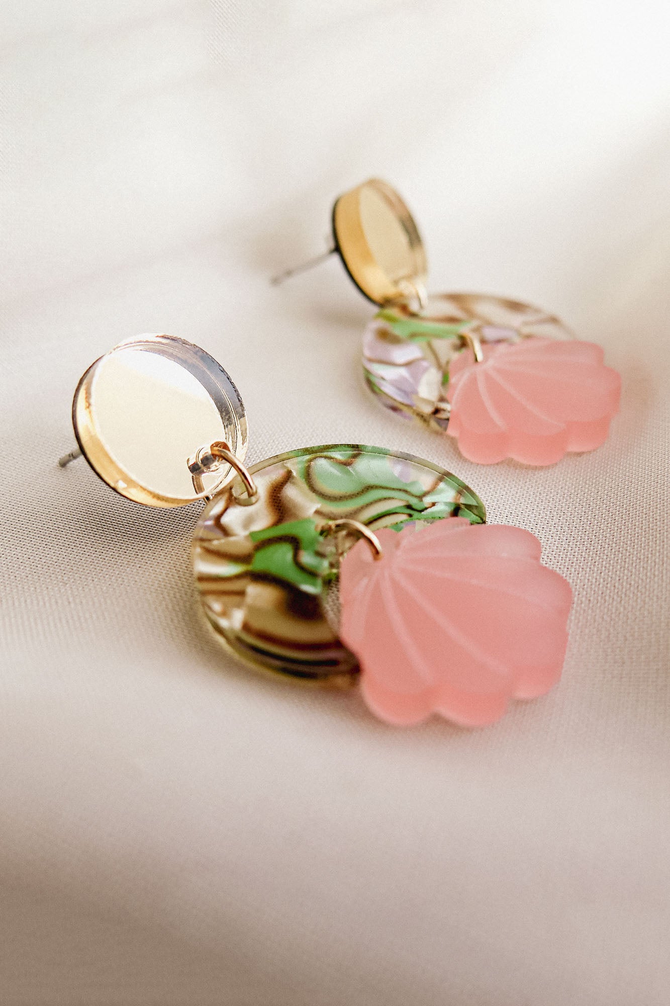 These handmade seashell earrings feature round gold studs, marbled green and cream discs, and pink translucent shell-shaped charms, all displayed on a soft beige fabric background. Lightweight and elegant for any occasion.
