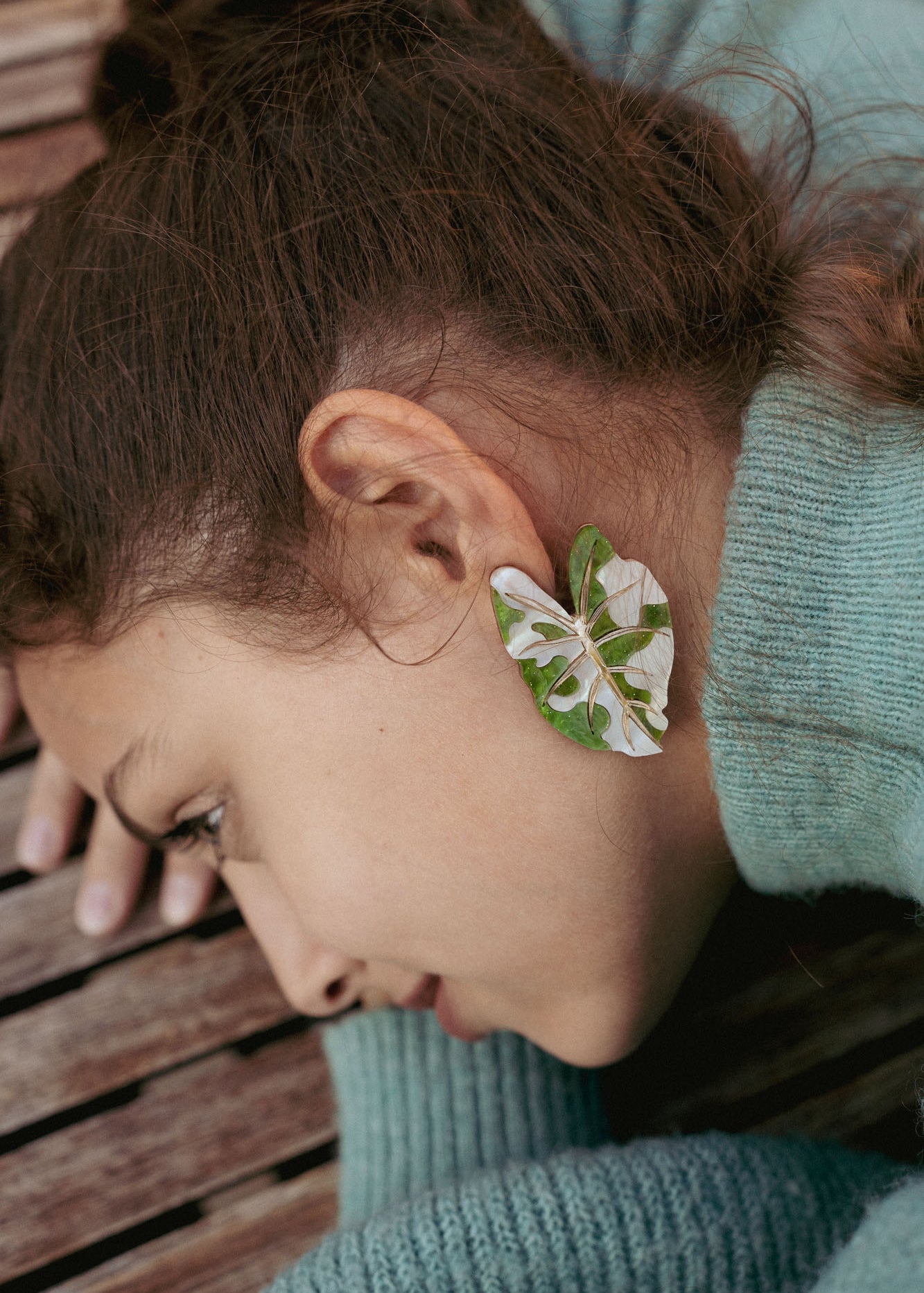 A person with brown hair lies on a wooden surface, wearing a green sweater. An Alocasia Earrings-inspired green and white leaf, reminiscent of hand-crafted jewelry, is placed over their ear, resembling an earring.
