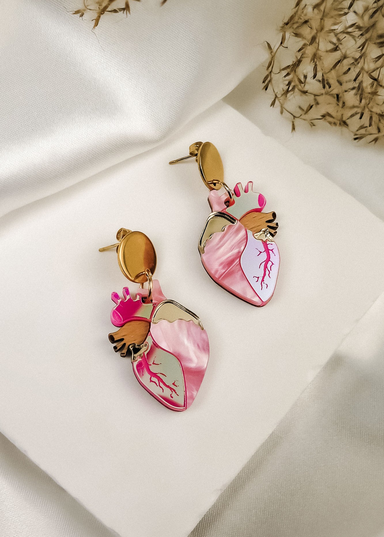 A pair of Anatomical Heart Earrings in a bold style, shaped like hearts with pink and gold details, are displayed on a white fabric surface with dried grass in the background.