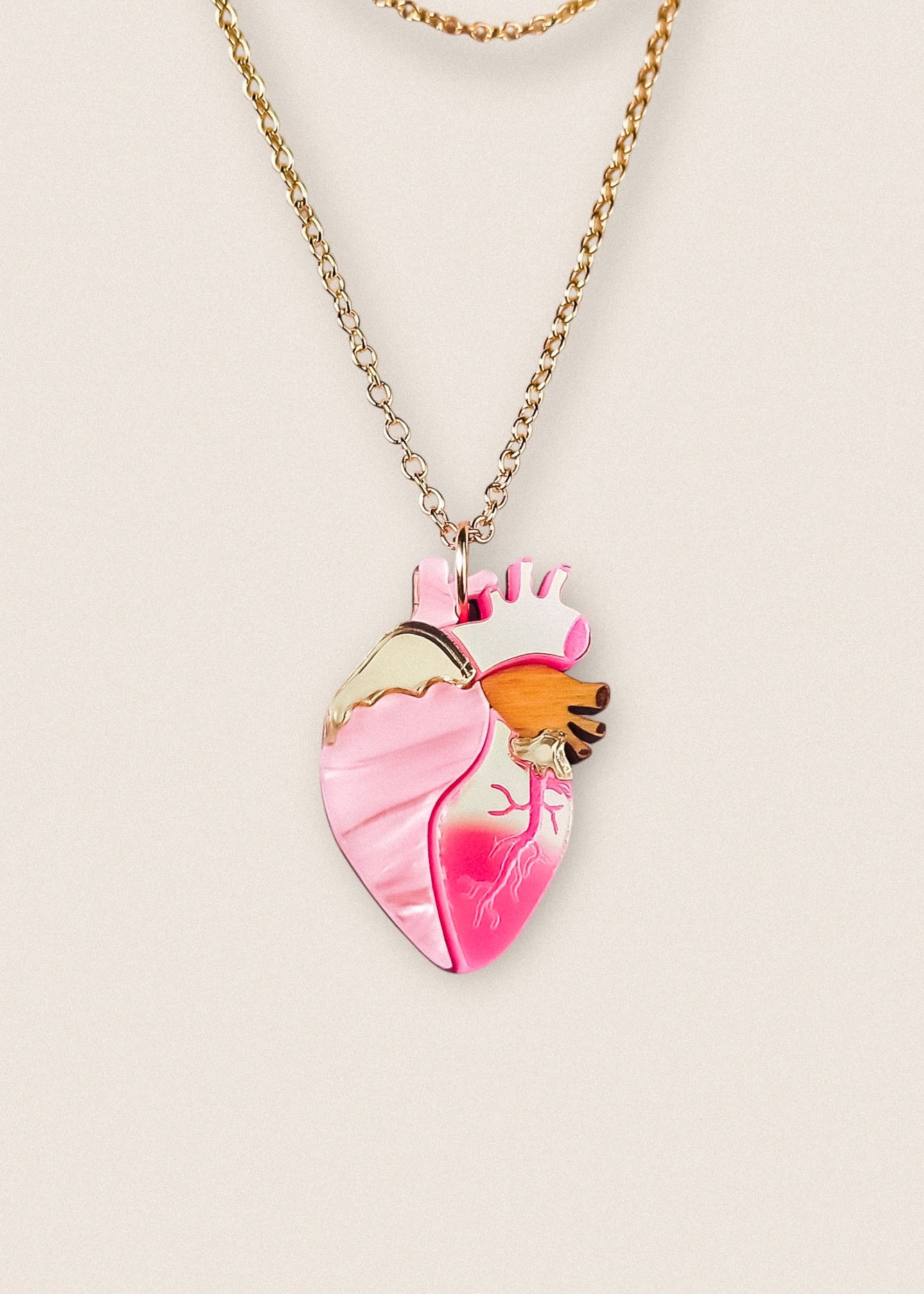 A gold chain anatomical heart necklace with a pink pendant featuring gold, orange, and dark pink details, displayed against a light beige background—perfect as a unique jewelry piece.