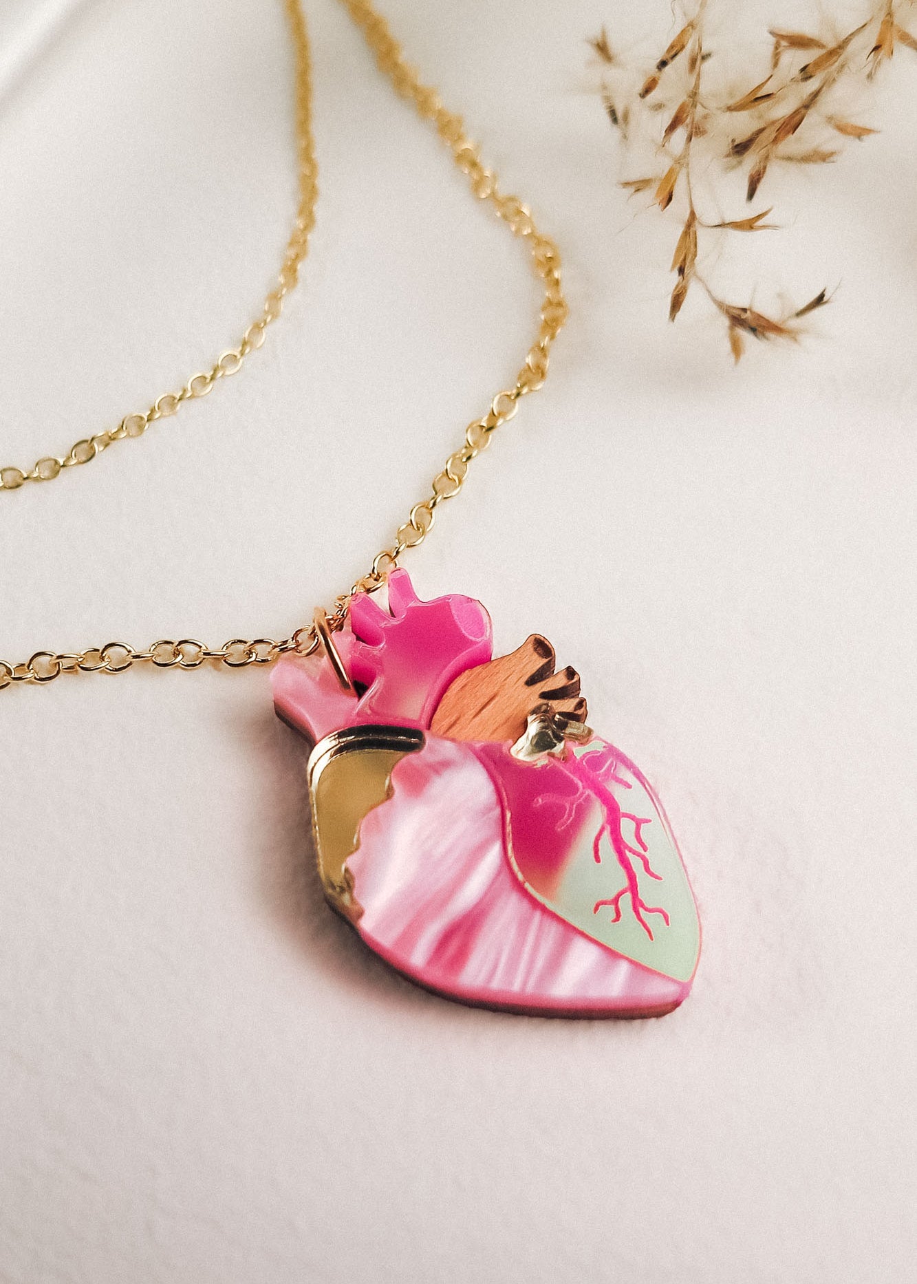 A gold chain anatomical heart necklace with a pendant in pink, mint green, and gold lies on a light surface, surrounded by dried grass—a unique jewelry piece for lovers of handcrafted jewelry.