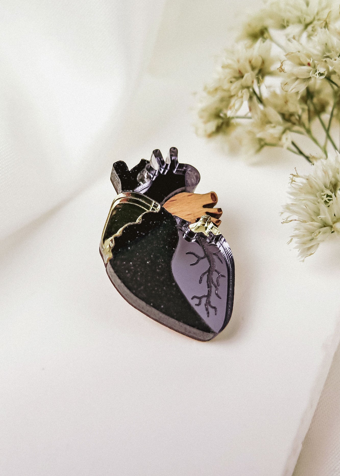 A handcrafted anatomical heart pin with black, purple, gold, and brown details rests on a white surface beside delicate white flowers—a striking statement piece for any collection.