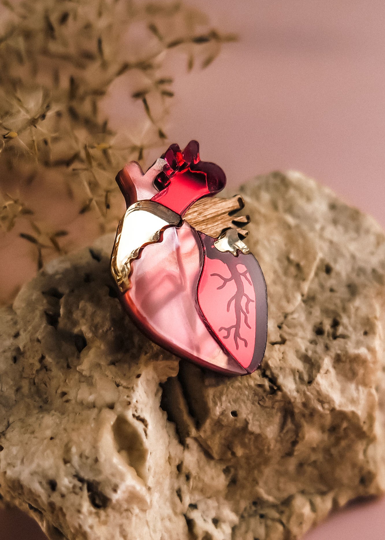 A handcrafted Anatomical Heart Pin, featuring red, pink, and gold details, rests on a textured beige rock with a blurred plant in the background—a true statement piece for any collection.