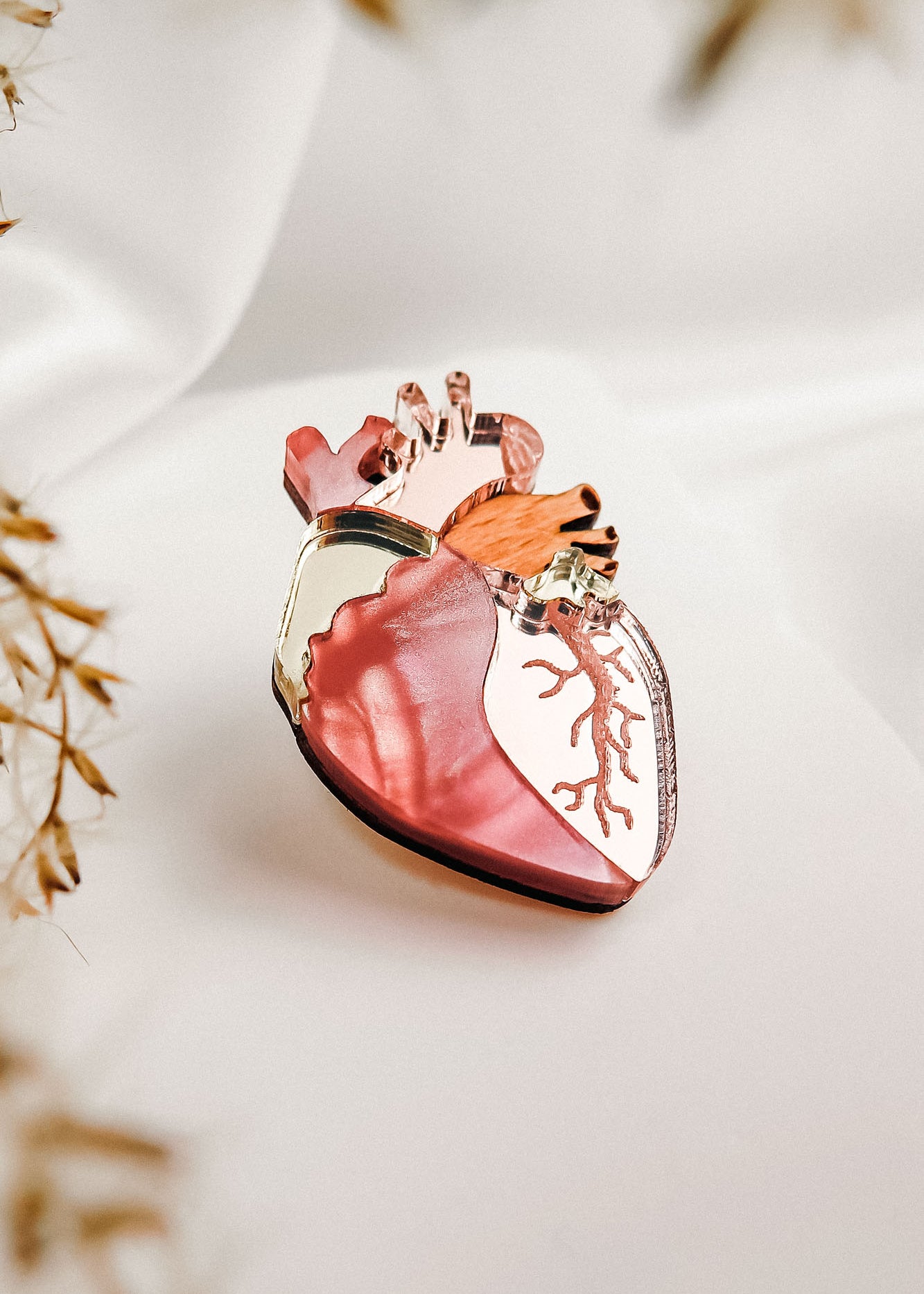 A unique handcrafted Anatomical Heart Pin, shaped with pink, red, gold, and copper hues, is displayed on soft white fabric with blurred dried plants in the foreground.