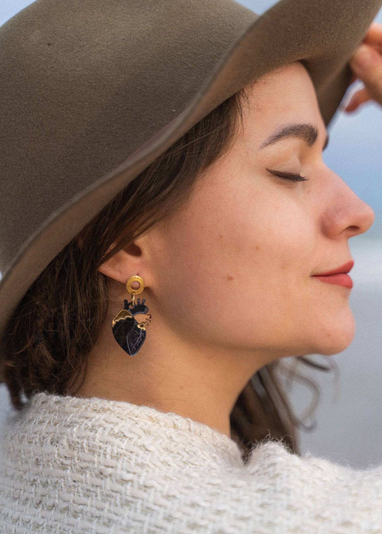 A woman in a brown hat and cream sweater smiles with her eyes closed, showing off handcrafted accessories—a black and gold leafy earring. The background is softly blurred.