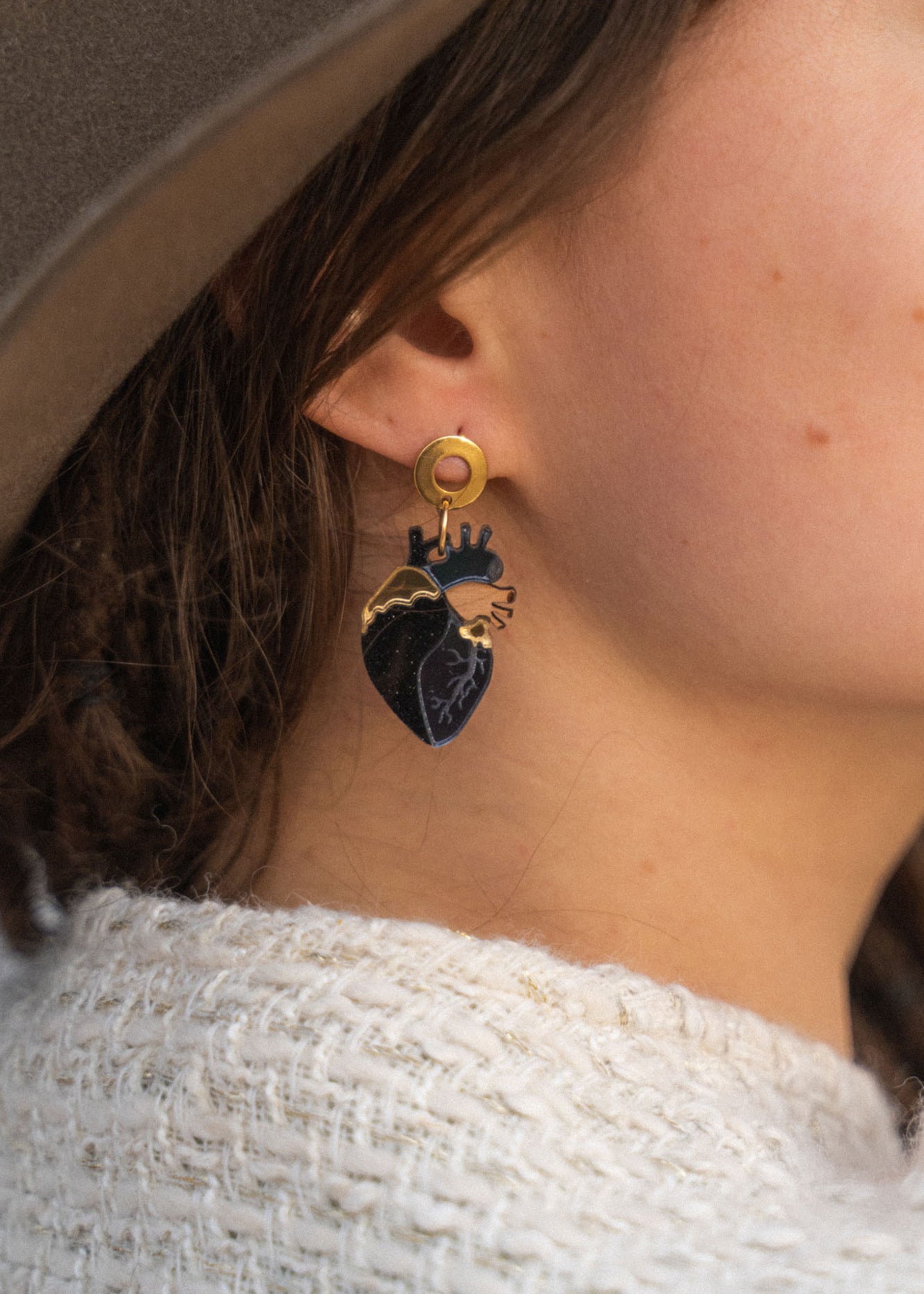 Close-up of a person wearing a gold and black Anatomical Heart Earring with gothic flair. The individual has light skin, brown hair, and is dressed in a beige hat and cream knitted sweater.