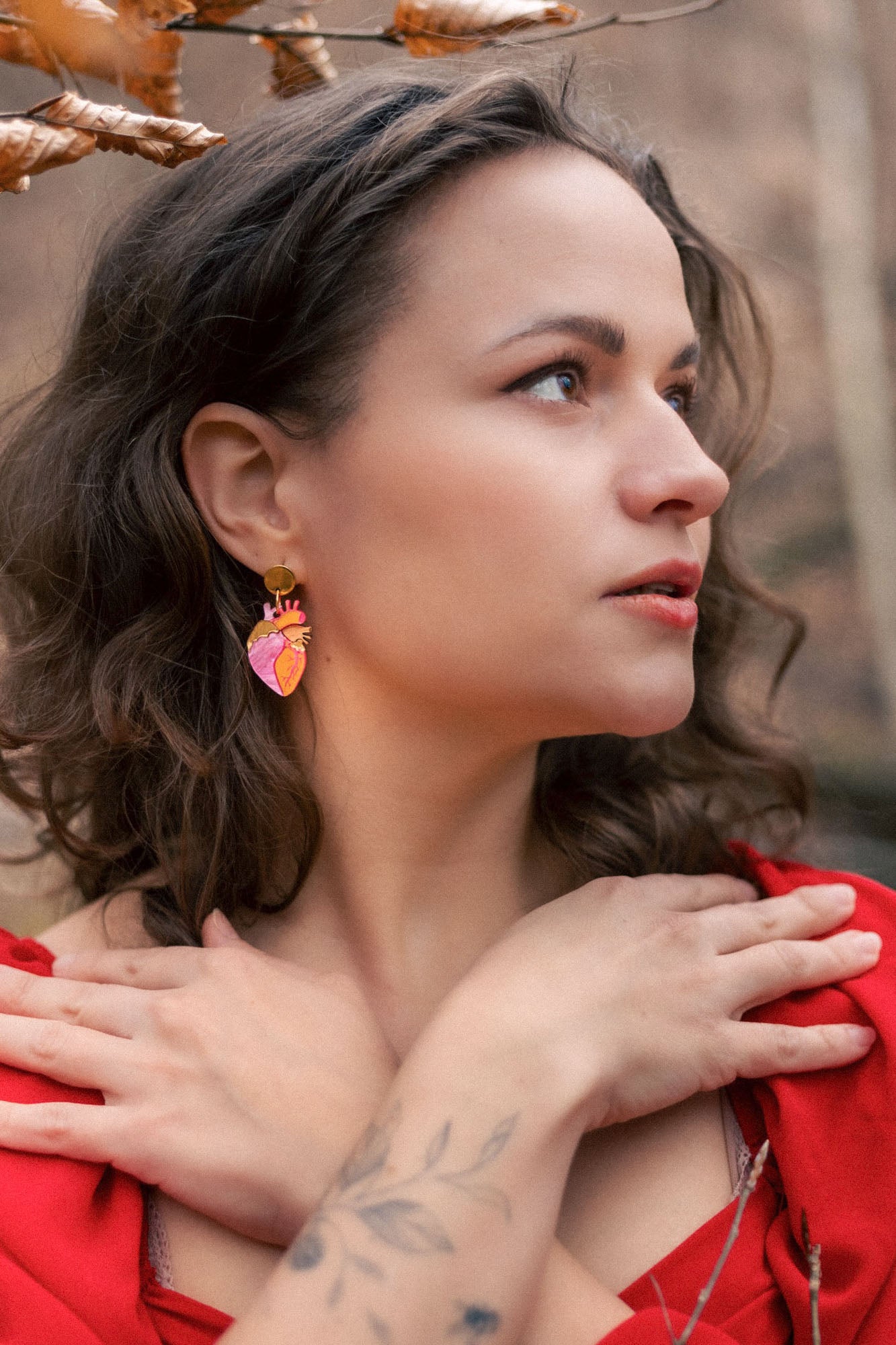 A woman with wavy brown hair and a tattooed forearm crosses her hands over her chest. She wears a red dress, bold style pink statement earrings, and stands outdoors among brown autumn leaves, looking to the side.