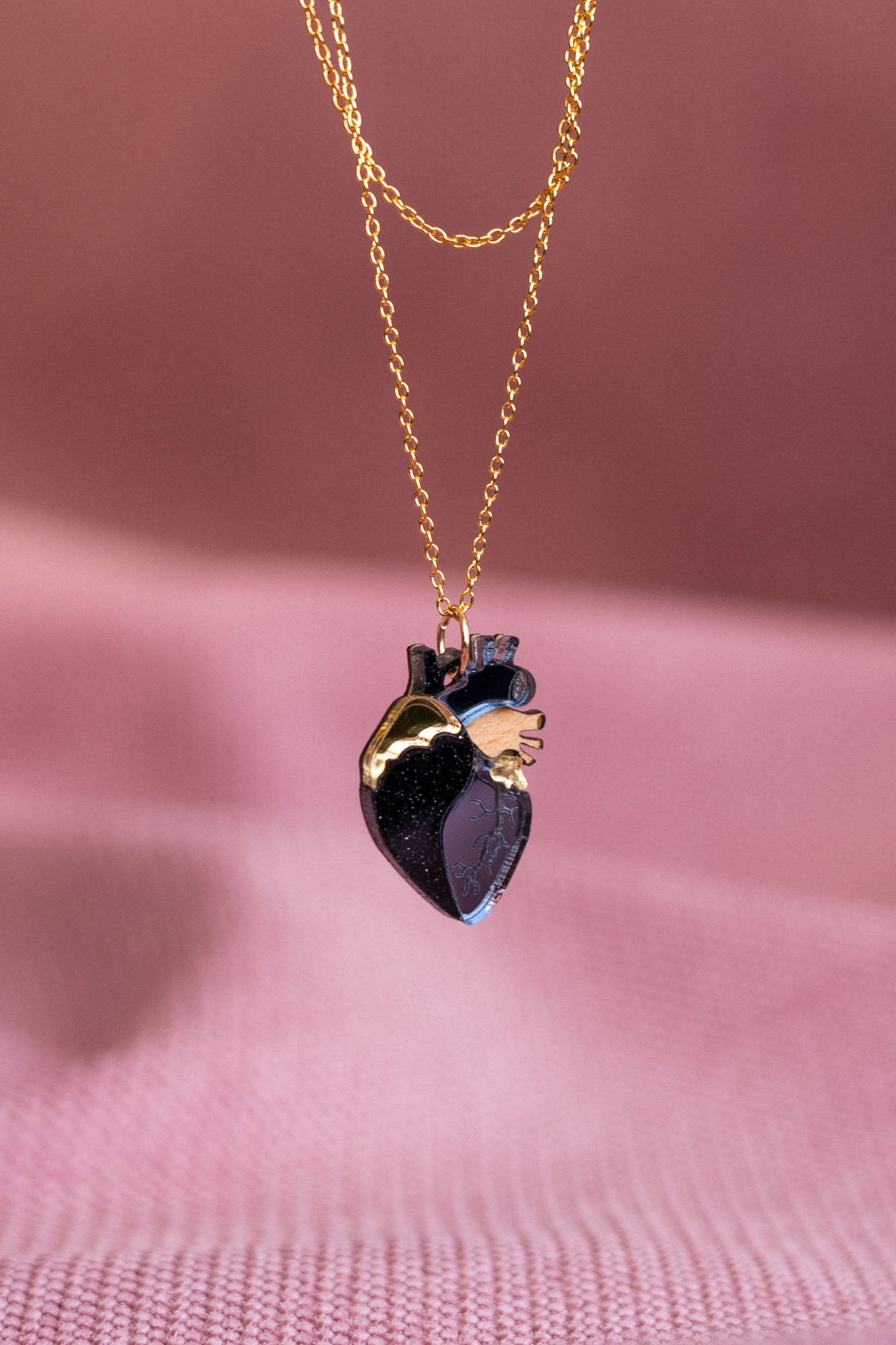 A handcrafted anatomical heart necklace with gold and dark purple details is captured against a soft pink textured background, highlighting this unique jewelry piece’s artistic design.