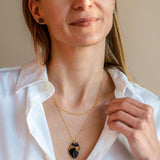 A woman with long light brown hair wears a white button-up shirt and a gold anatomical heart necklace, looking slightly to the side with a soft smile against a plain beige background.