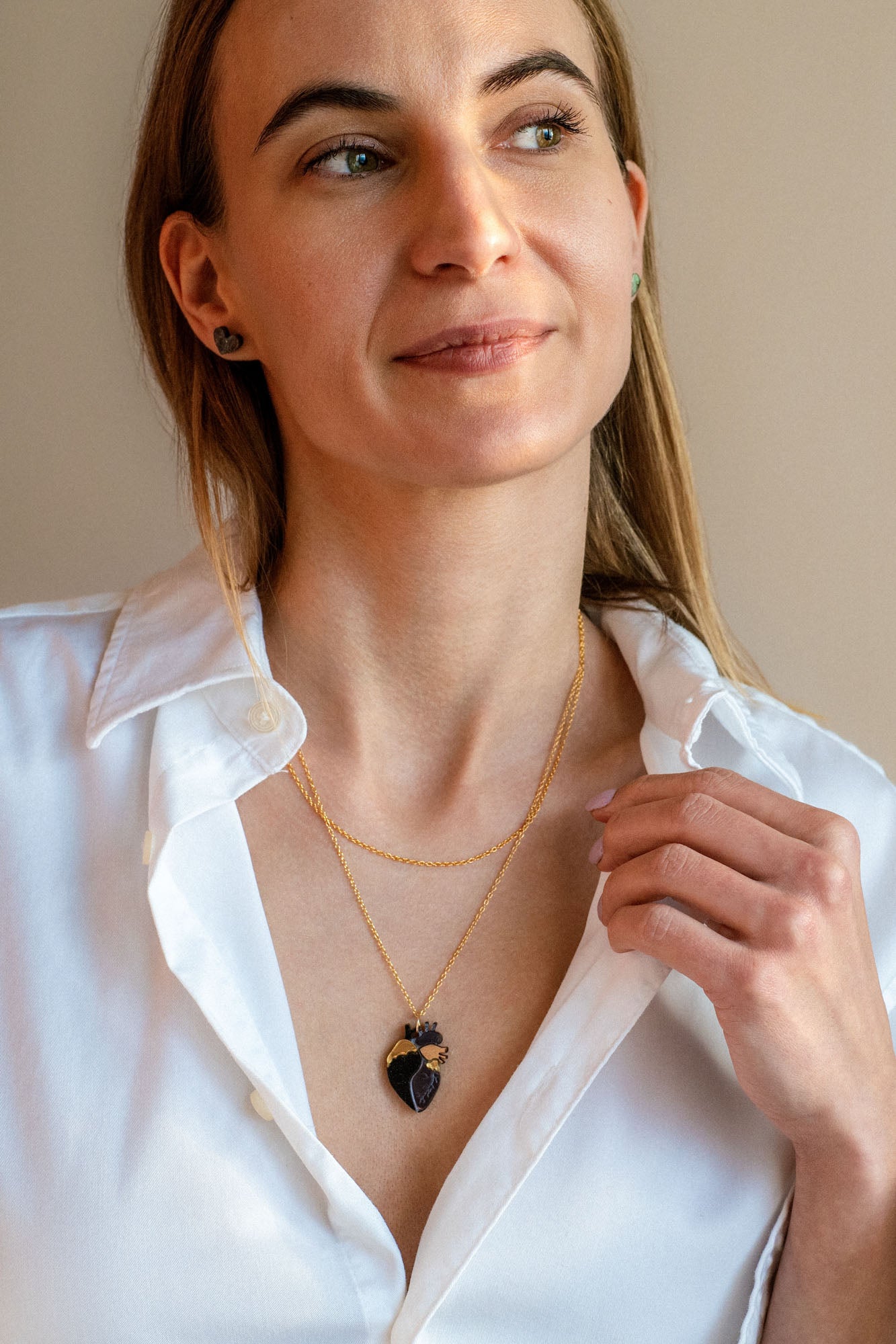A woman with long light brown hair wears a white button-up shirt and a gold anatomical heart necklace, looking slightly to the side with a soft smile against a plain beige background.