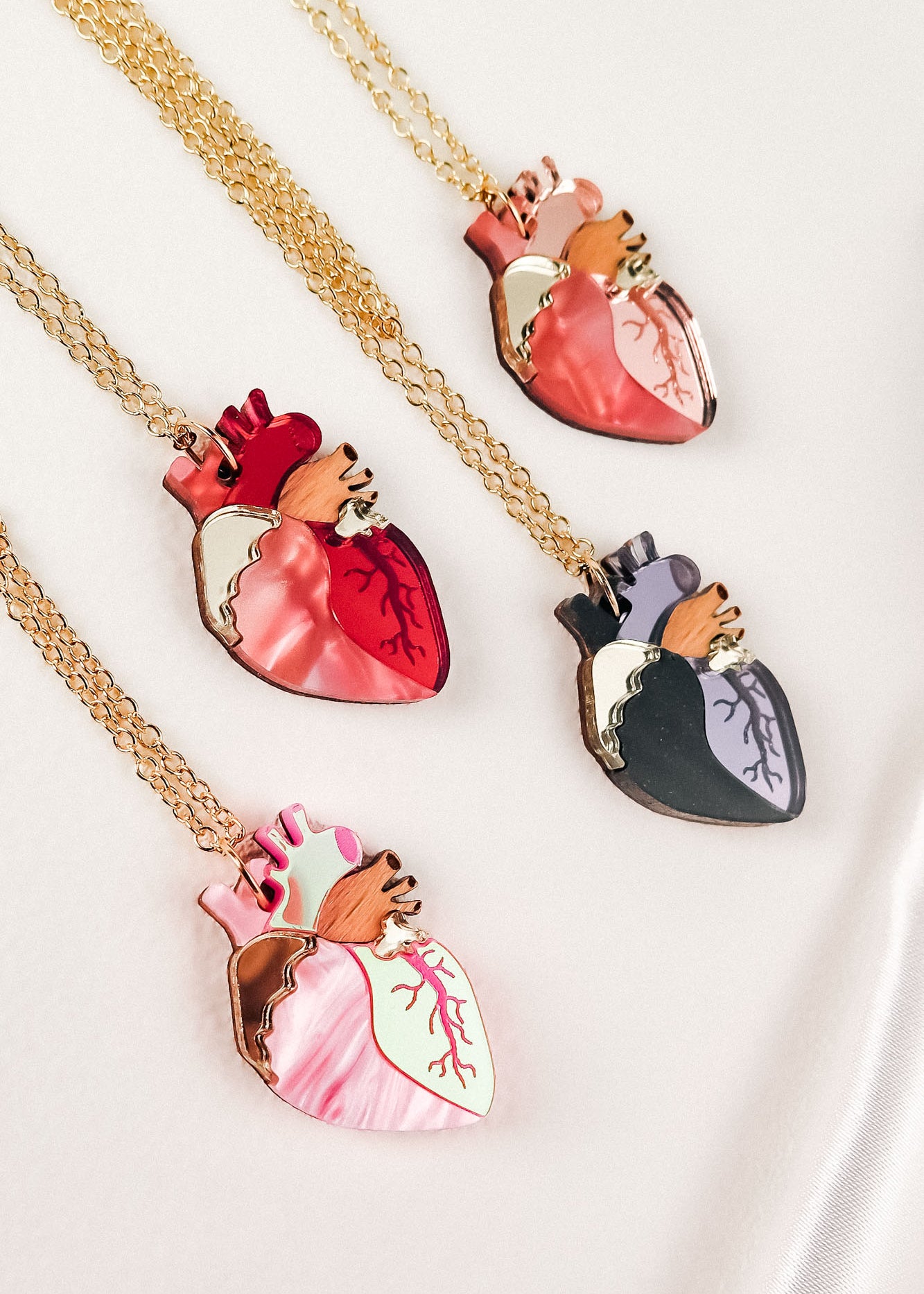Four handcrafted jewelry necklaces with gold chains, each featuring an anatomical heart necklace pendant in dark red, pink, black and purple, or pink and mint green—making each one a unique jewelry piece displayed on a light background.