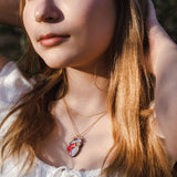 A young woman with long light brown hair wearing a white top and layered gold necklaces, including a unique anatomical heart necklace, stands outdoors with sunlight on her face and hands behind her head.