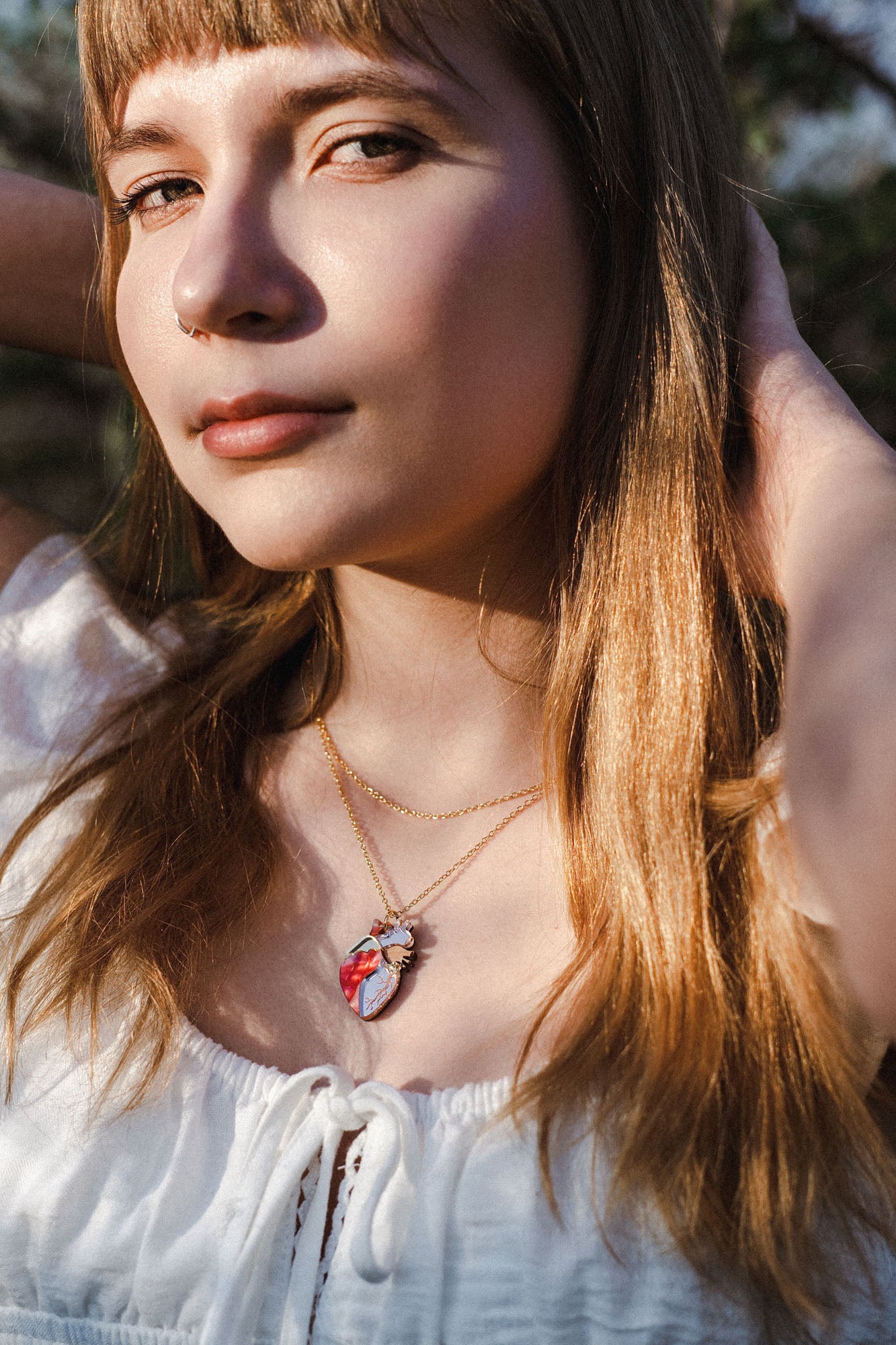 A young woman with long light brown hair wearing a white top and layered gold necklaces, including a unique anatomical heart necklace, stands outdoors with sunlight on her face and hands behind her head.