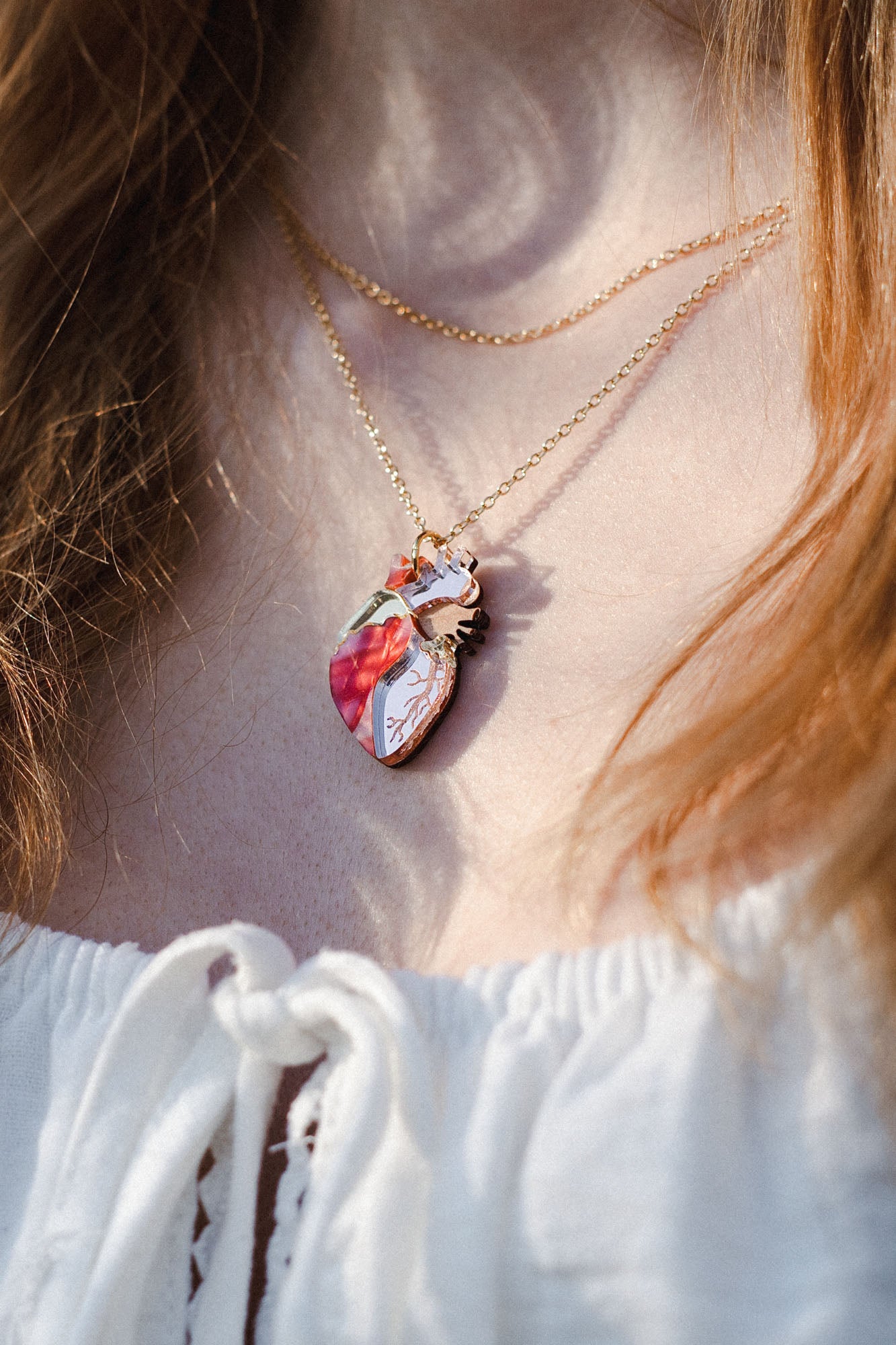Close-up of a person wearing an anatomical heart necklace in red, pink, and white—a striking piece of unique jewelry. The handcrafted pendant hangs over a white top with a lace-up detail.