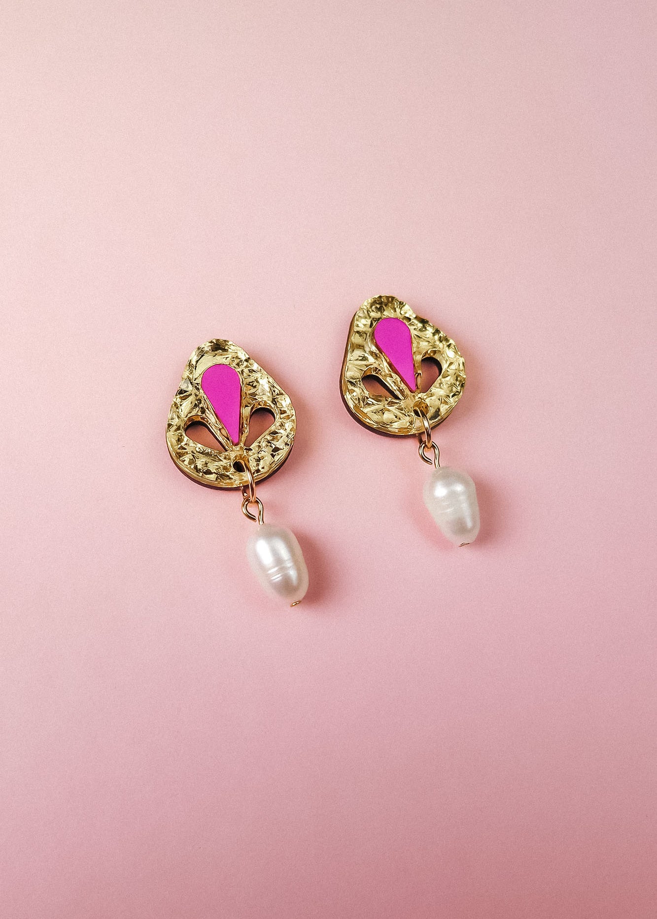 A pair of handcrafted gold-toned drop earrings with pink accents and a dangling white pearl, displayed on a soft pink background.