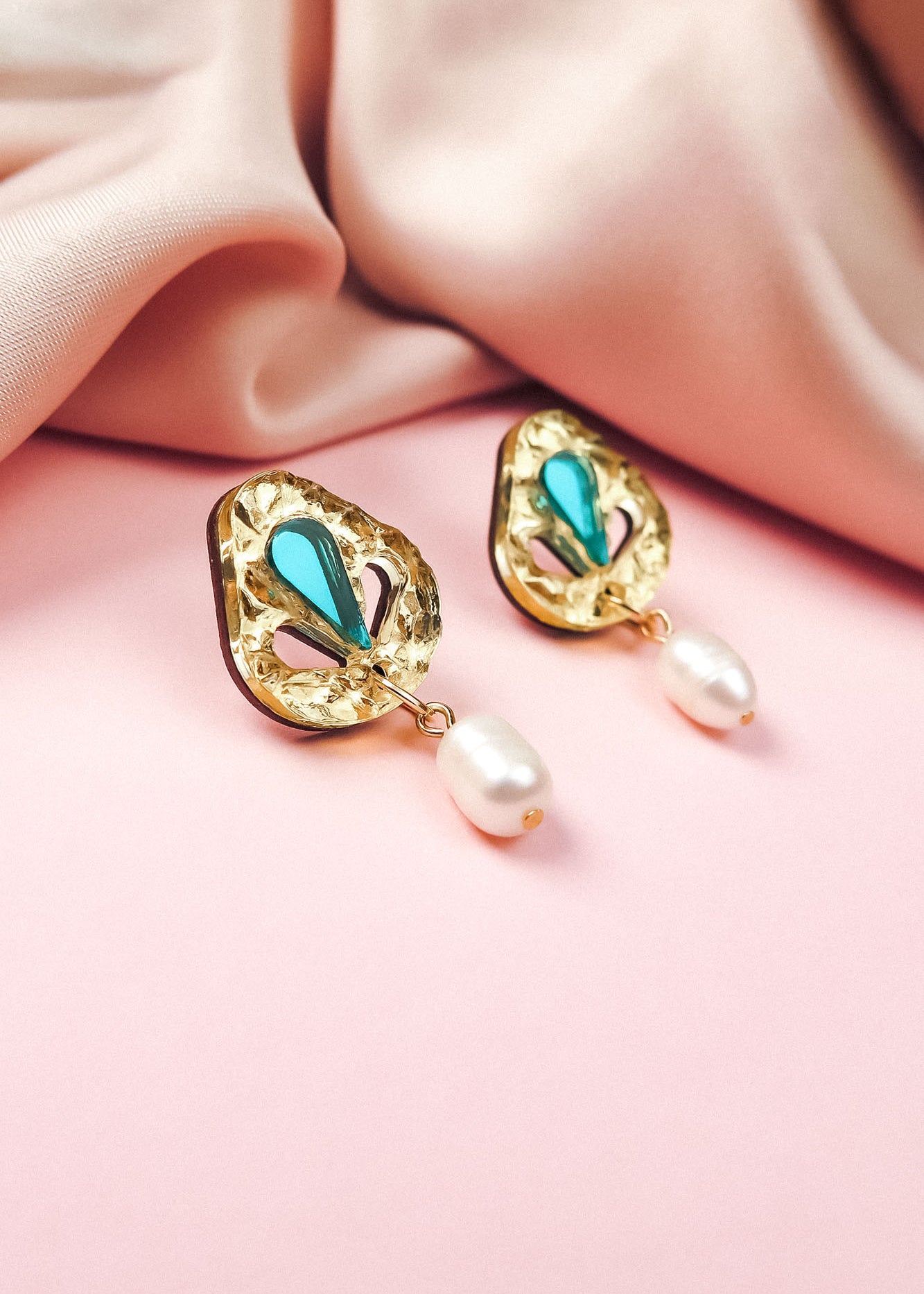 A pair of handcrafted gold earrings with turquoise teardrop accents and dangling white pearls, perfect for minimalist fashion, displayed on a soft pink surface beside a folded blush-colored fabric.
