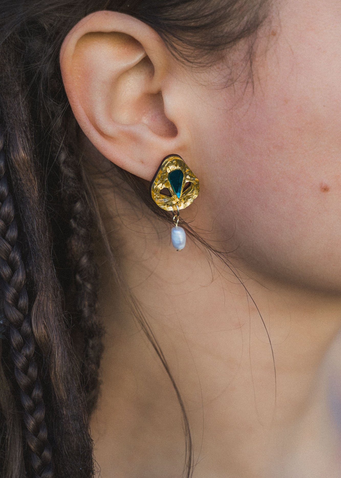 Close-up of a person’s ear wearing handcrafted Basic Drop Earrings featuring a gold setting with a green gemstone and dangling white pearl; the person has braided brown hair, perfect for minimalist fashion lovers.