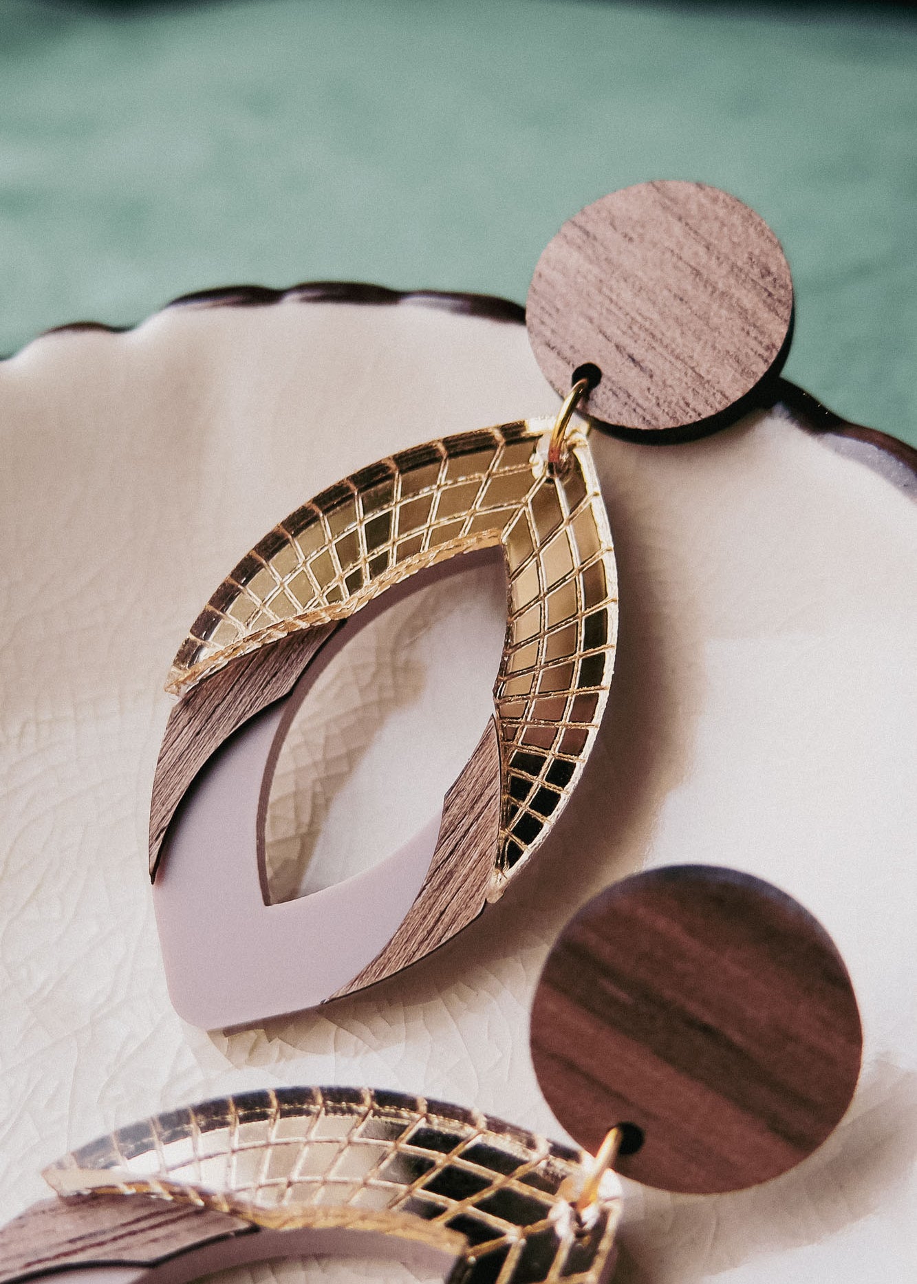 Close-up of handcrafted earrings with circular wooden studs and geometric drop pendants featuring gold lattice, wood accents, and muted purple elements, displayed on a cream-colored ceramic dish. Made with premium acrylic glass details.