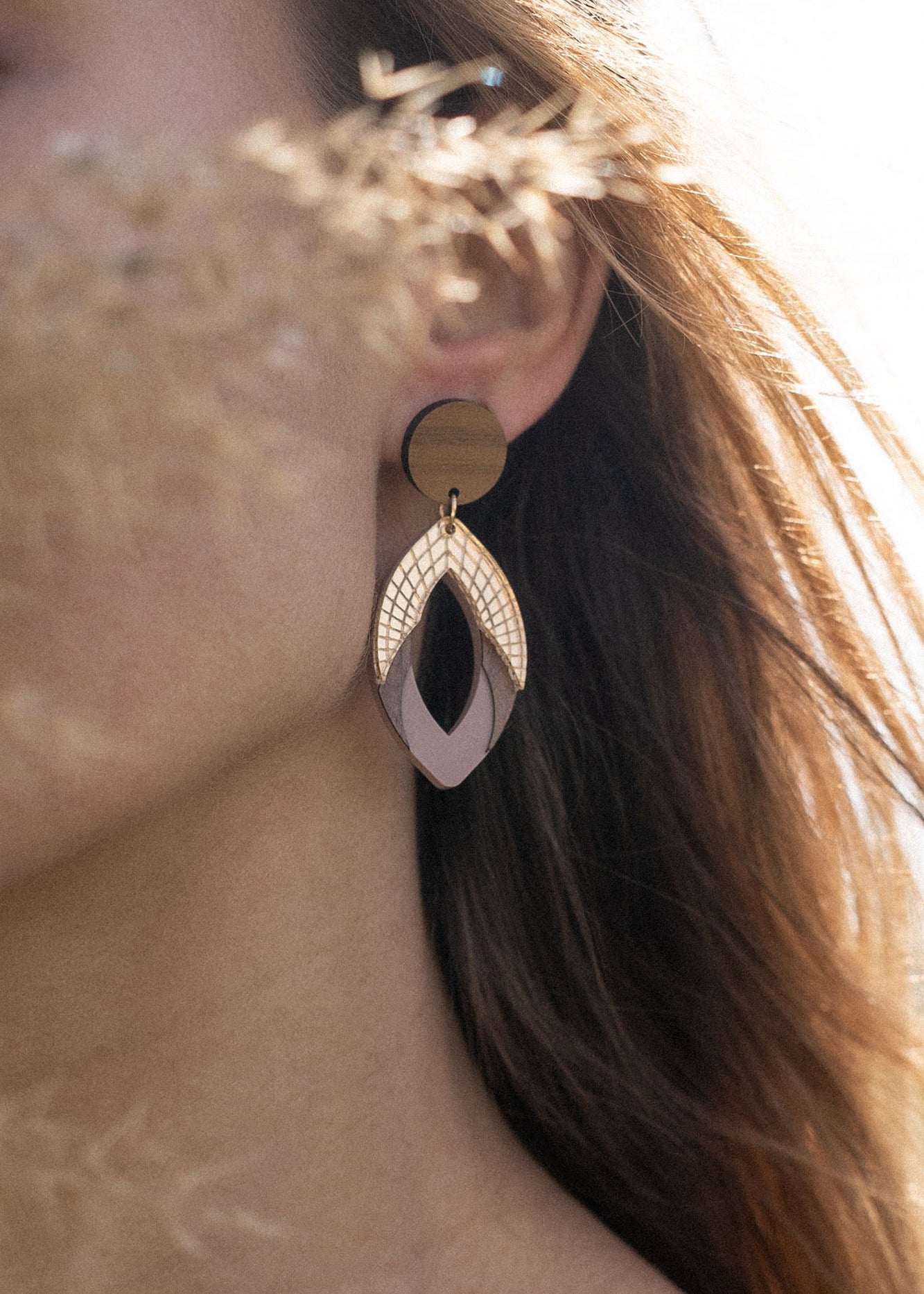 A woman with long hair wearing a mauve top and large, handcrafted bear earrings stands outdoors. Soft sunlight illuminates her face, partially obscured by dried grass in the foreground.
