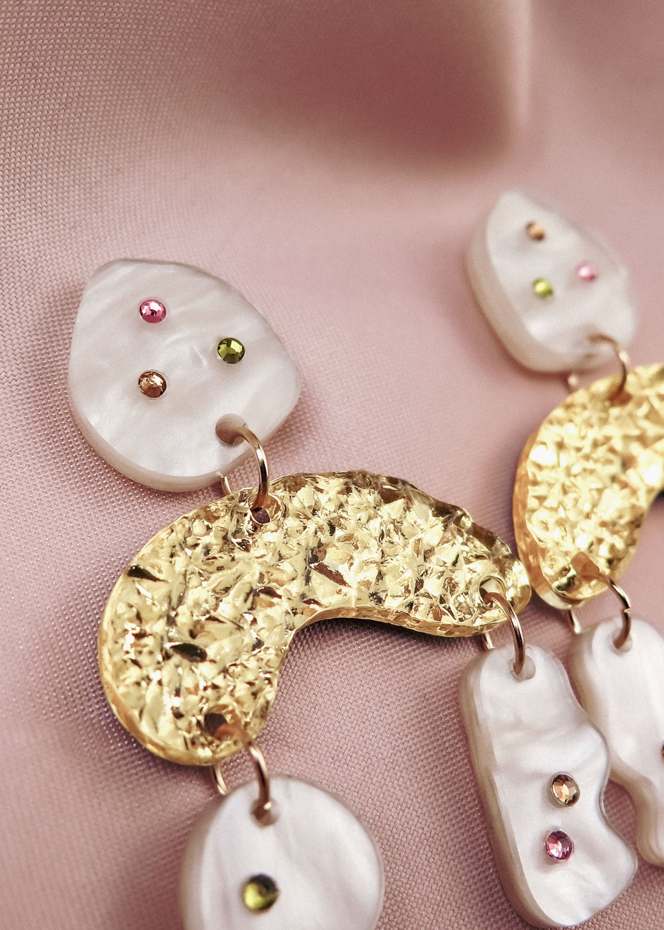 Close-up of abstract dangle earrings with textured gold shapes and marbled white pieces, decorated with small pink, green, and amber rhinestones, displayed on soft, light pink fabric.