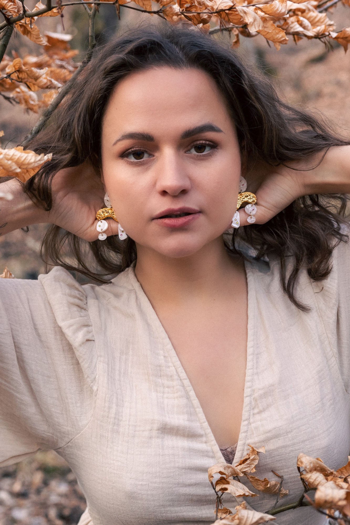 A woman with wavy brown hair poses outdoors among dry autumn leaves, wearing a light beige dress and boho chic dangle earrings adorned with rhinestones, her hands touching her hair and an intense, confident expression on her face.