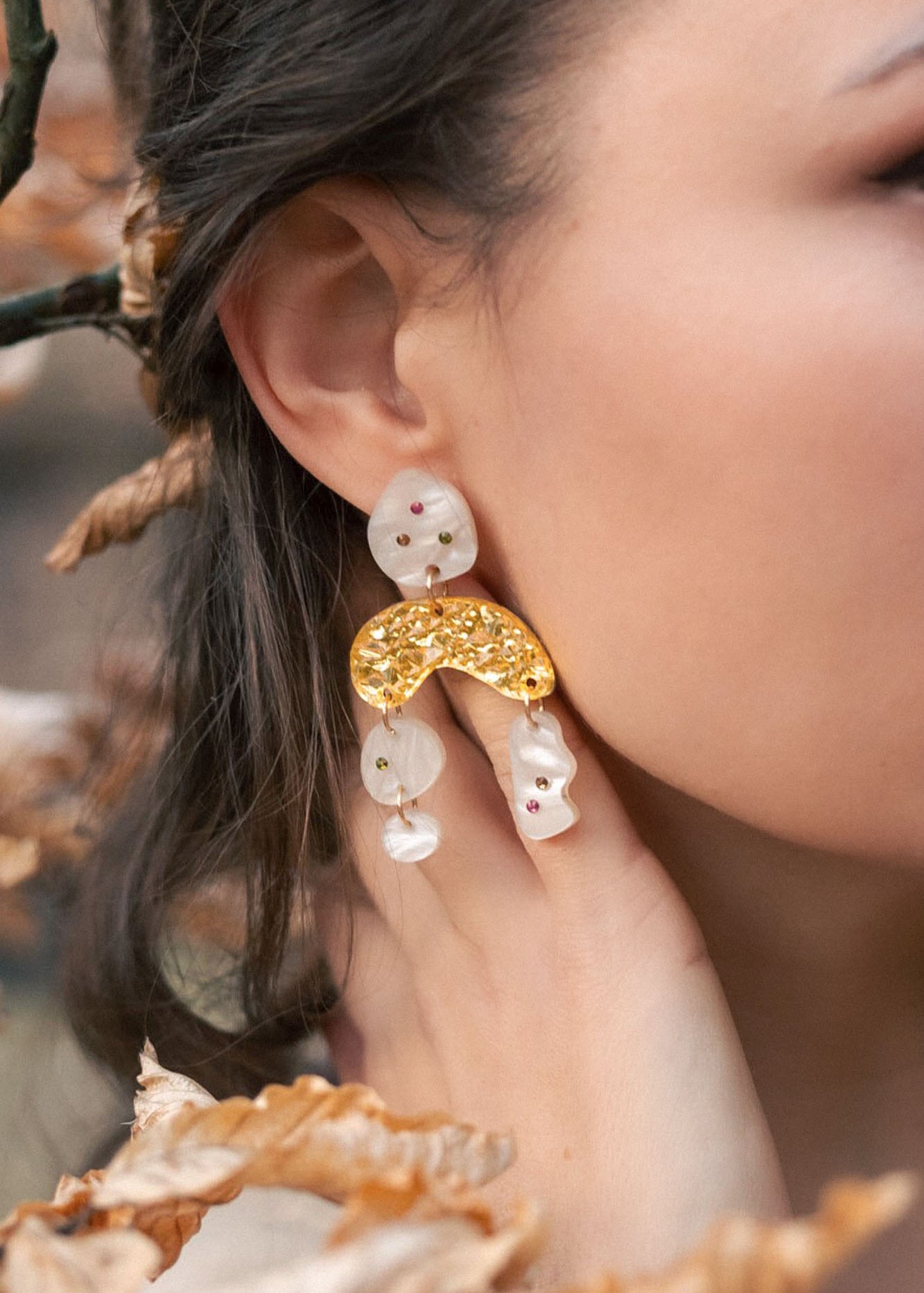 A close-up of a womans ear wearing unique boho chic dangle earrings with gold, pearl-like abstract shapes, and subtle rhinestones. Her hand touches her neck, surrounded by brown autumn leaves and branches.