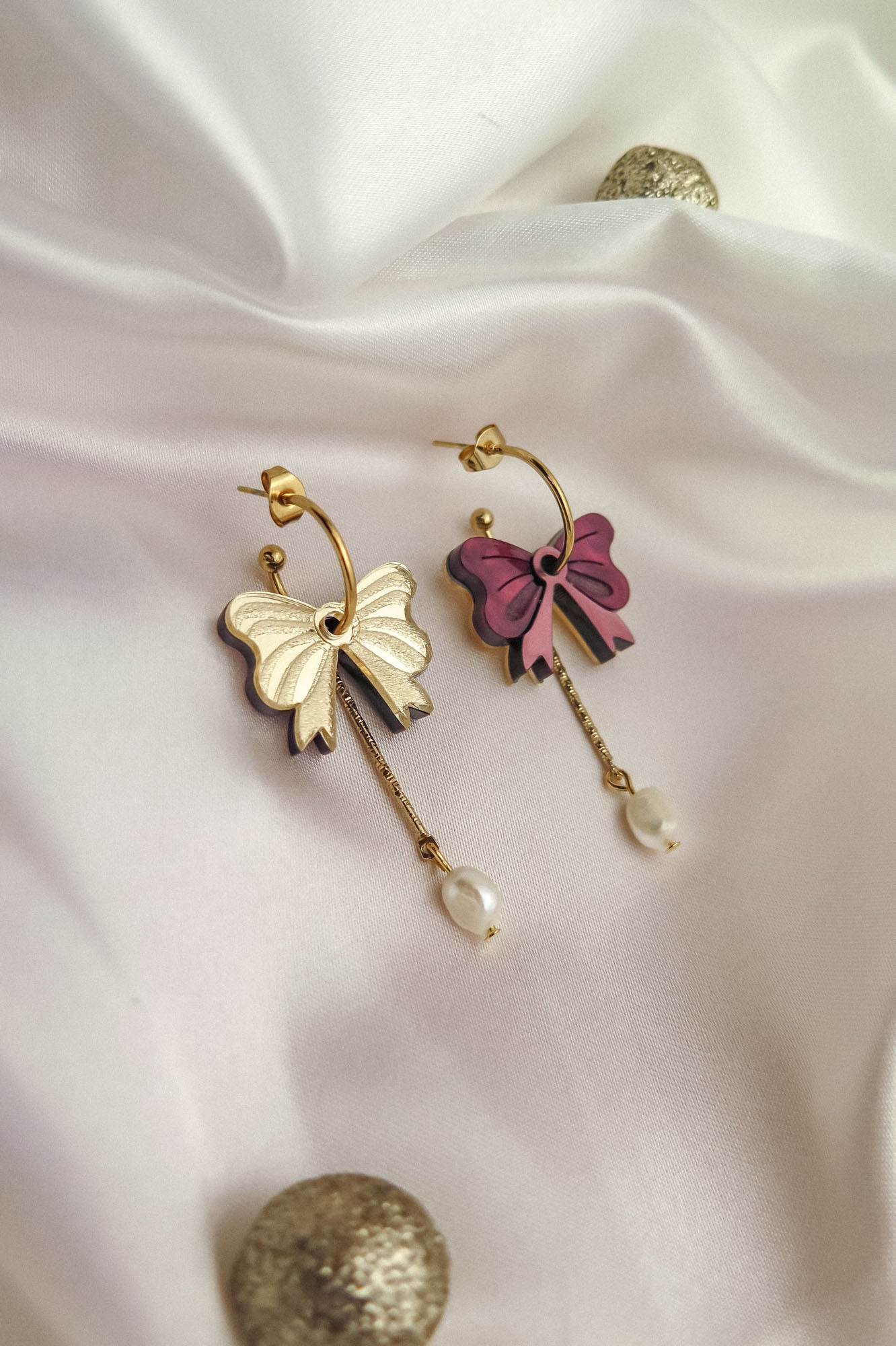 Two gold-plated hoop earrings with bow-shaped charms—one cream and one pink—each with a dangling gold chain and a freshwater pearl, displayed on white fabric with decorative gold spheres.