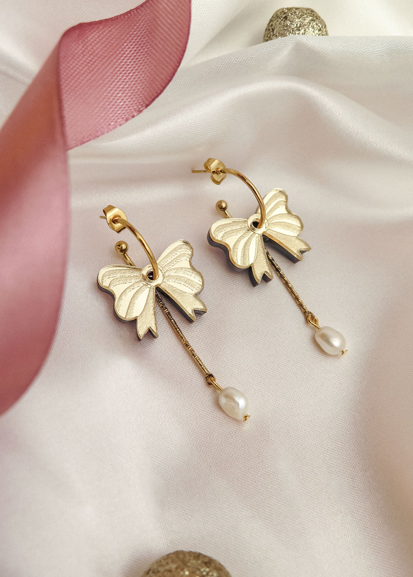 A pair of gold-plated earrings shaped like bows with dangling freshwater pearl accents, displayed on white satin fabric with a pink ribbon and gold decorative balls nearby.