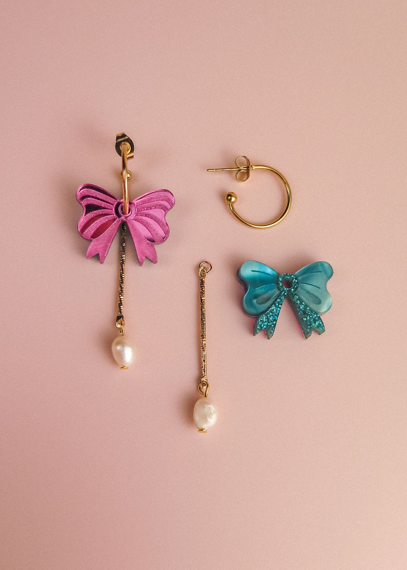 Three mismatched interchangeable dangle earrings on a pink surface: a gold-plated c-hoop, a dangling earring with a pink bow and freshwater pearl, and another with a blue bow, sequins, and pearl.