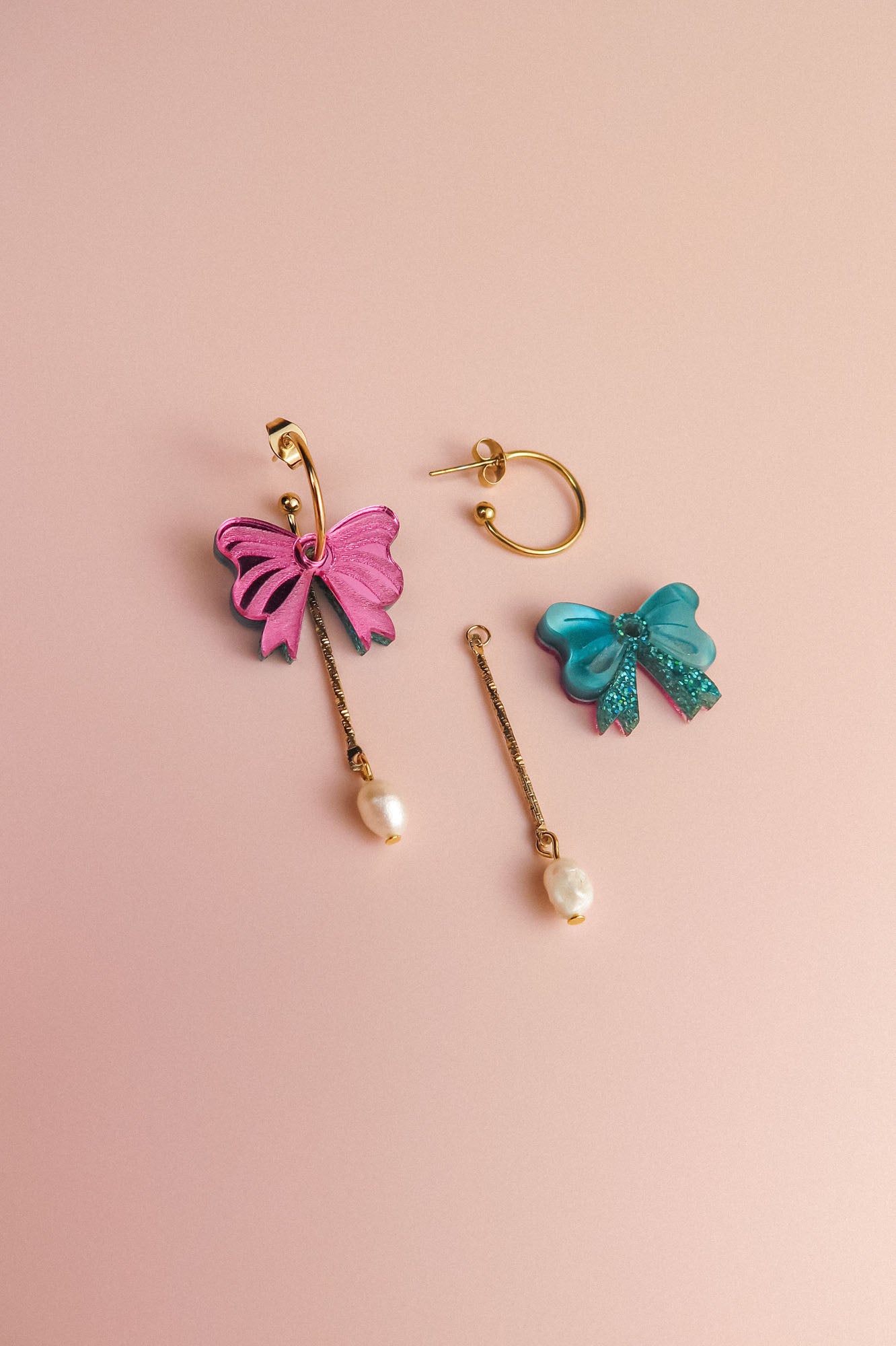 A pair of mismatched interchangeable dangle earrings on a pink background: one earring features a pink bow with a dangling freshwater pearl, while the other has a blue bow with a gold-plated c-hoop and bar, also adorned with a pearl.