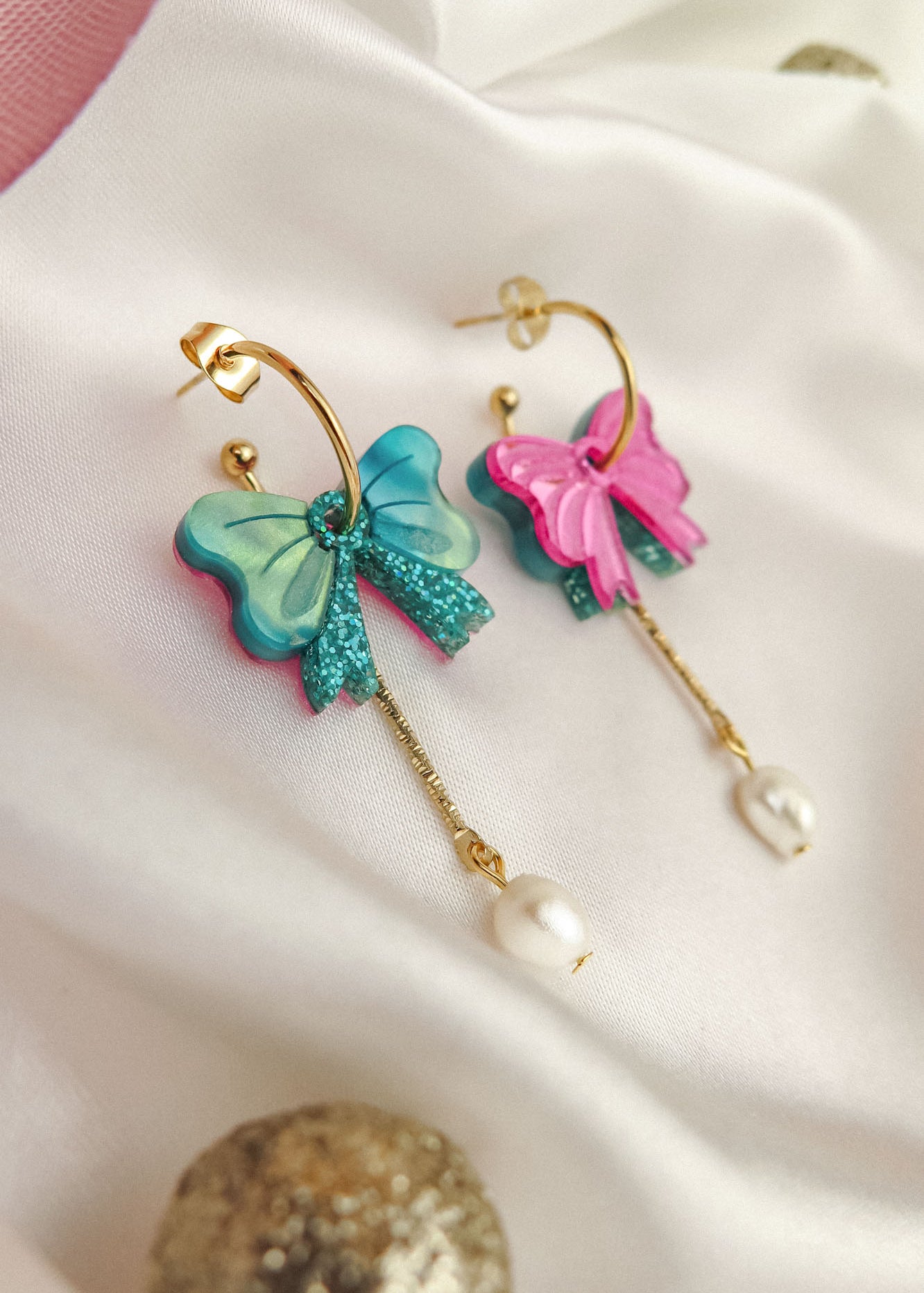 Gold-plated c-hoop earrings with colorful butterfly charms—one blue and one pink—feature freshwater pearl and gold chain accents. These interchangeable dangle earrings are displayed on a white fabric background.